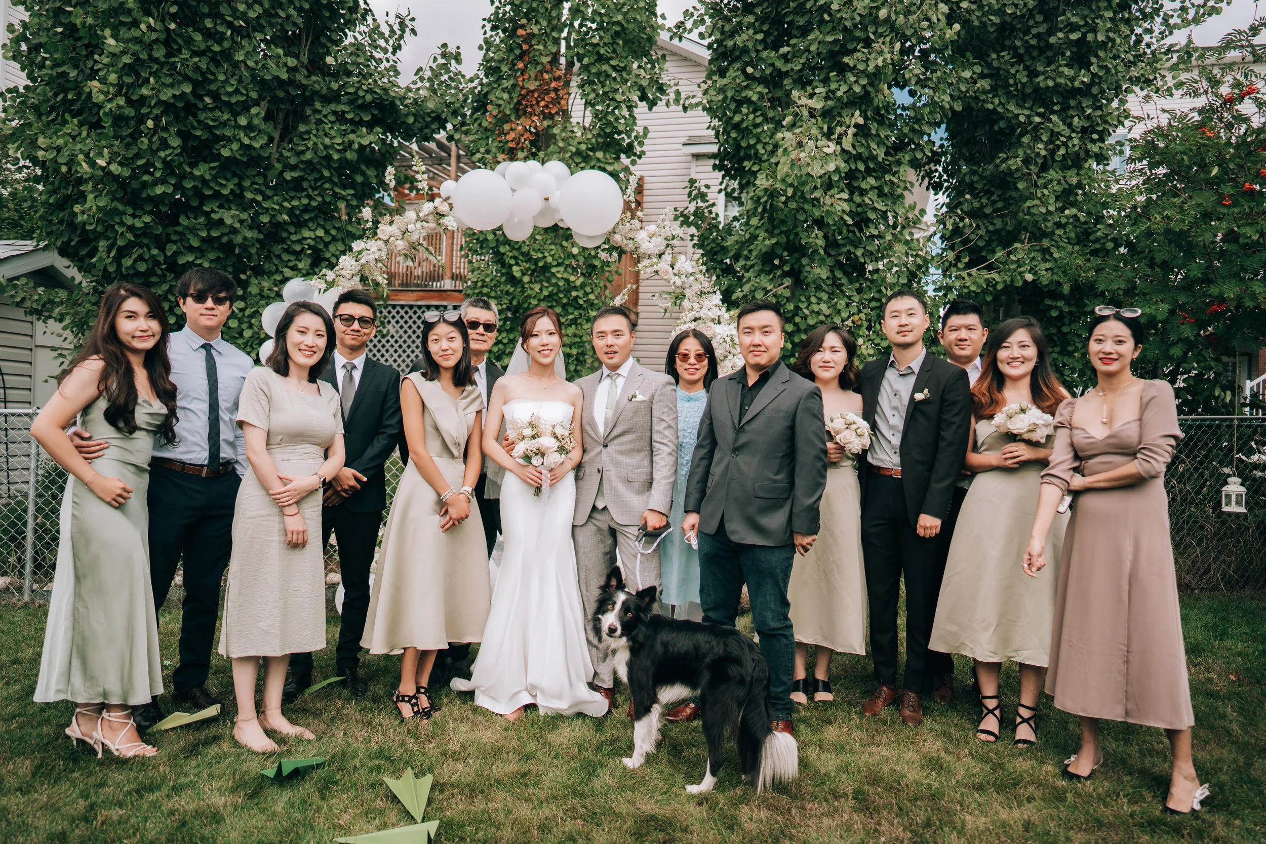 Group of people gathered outdoors for a wedding celebration, with a bride in a white gown and groom in a gray suit at the center, surrounded by family and friends, some holding bouquets and a dog in front.