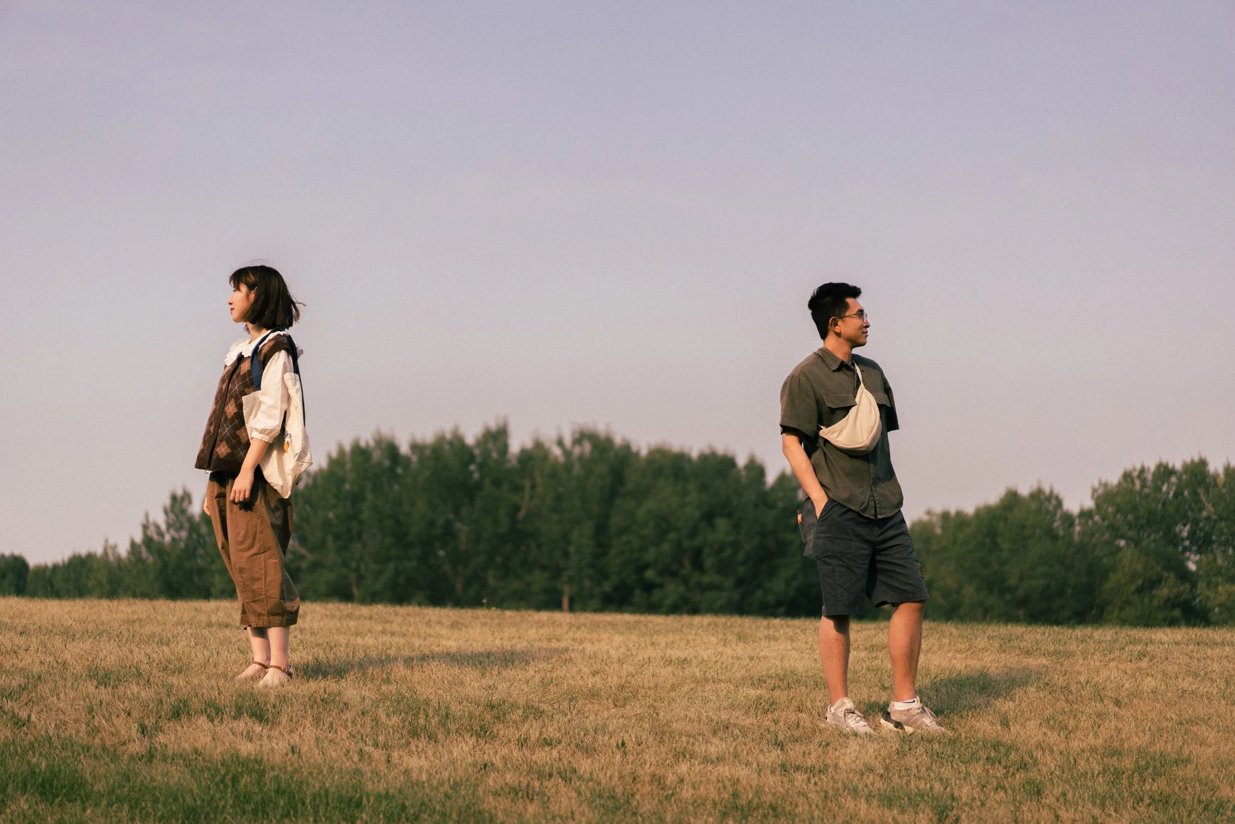A young woman and a young man standing apart on a grassy field with trees in the background, looking in opposite directions during sunset.