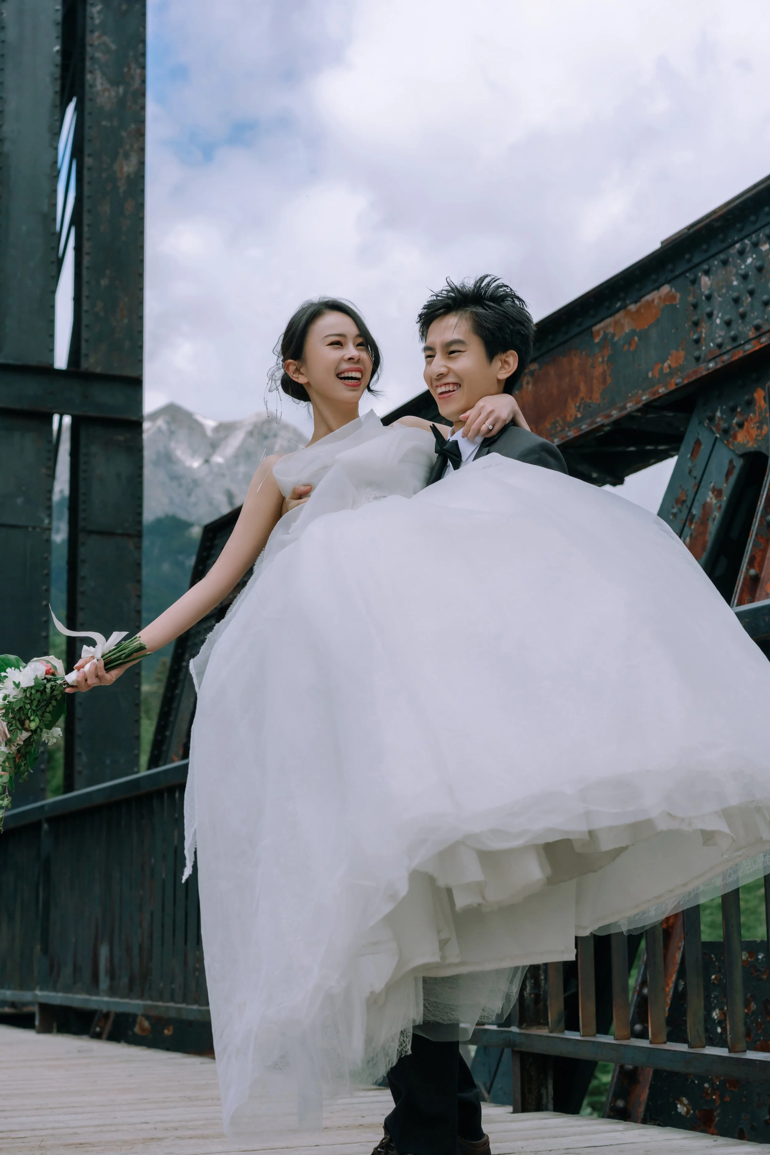 A bride and groom smiling as the groom lifts the bride on a bridge with a mountain and cloudy sky background.