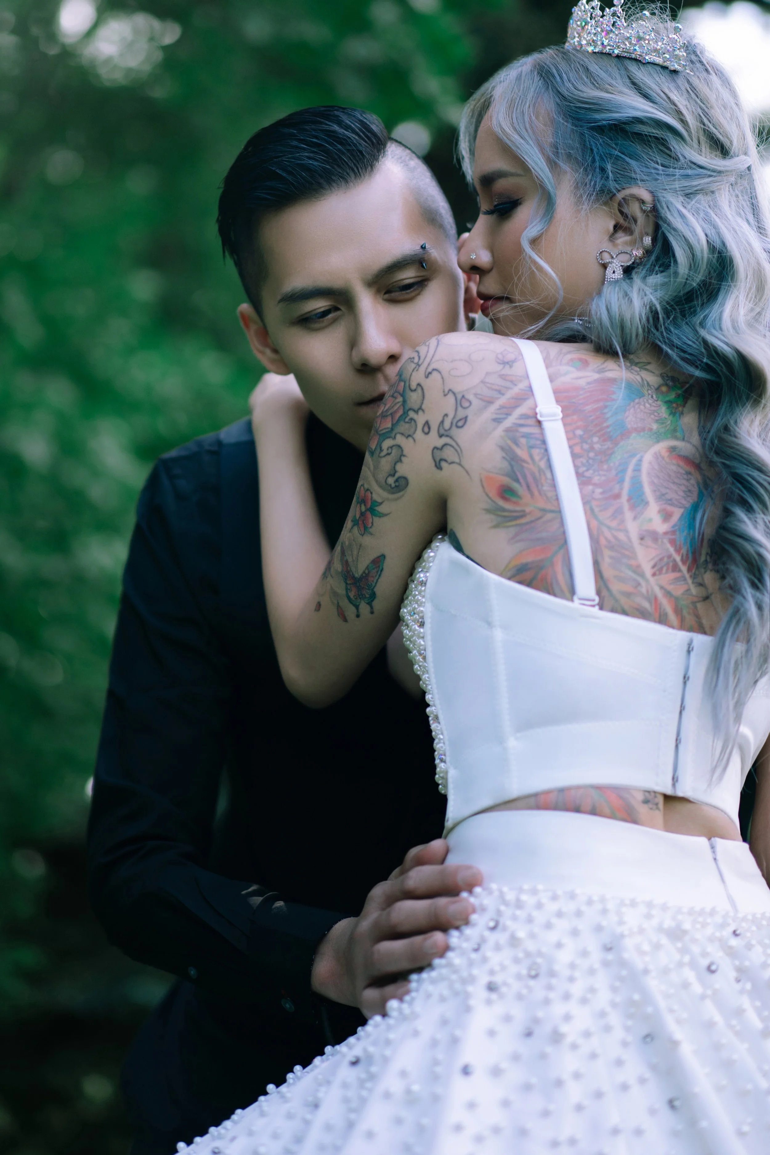 A couple embraces outdoors, the woman wearing a white, pearl and beaded dress with tattoos on her arm and back, a tiara, and silver hair, while the man wears a black suit with short hair and facial piercings.