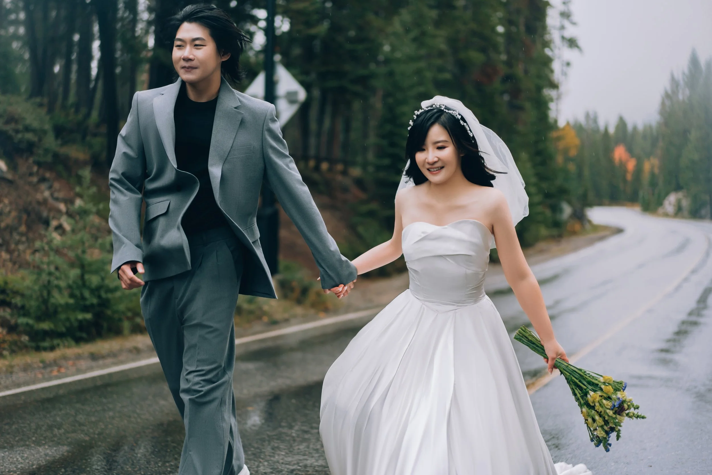 A couple in wedding attire walking hand in hand along a wet road in a forested area, with the woman holding a bouquet of yellow and purple flowers.