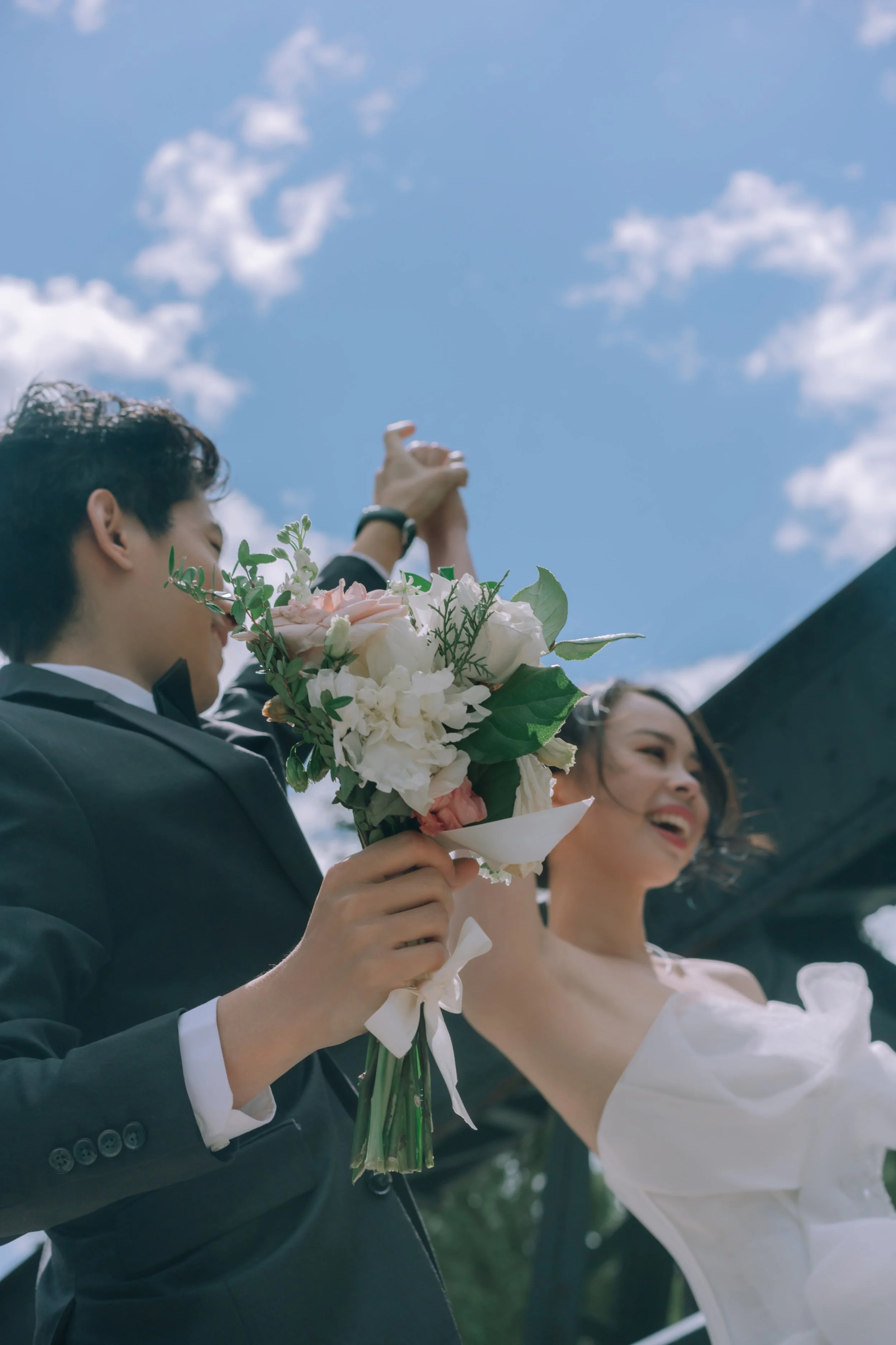 A newlywed couple celebrating outdoors under a blue sky with clouds; the groom holding a bouquet of flowers and the bride smiling with her arm raised.