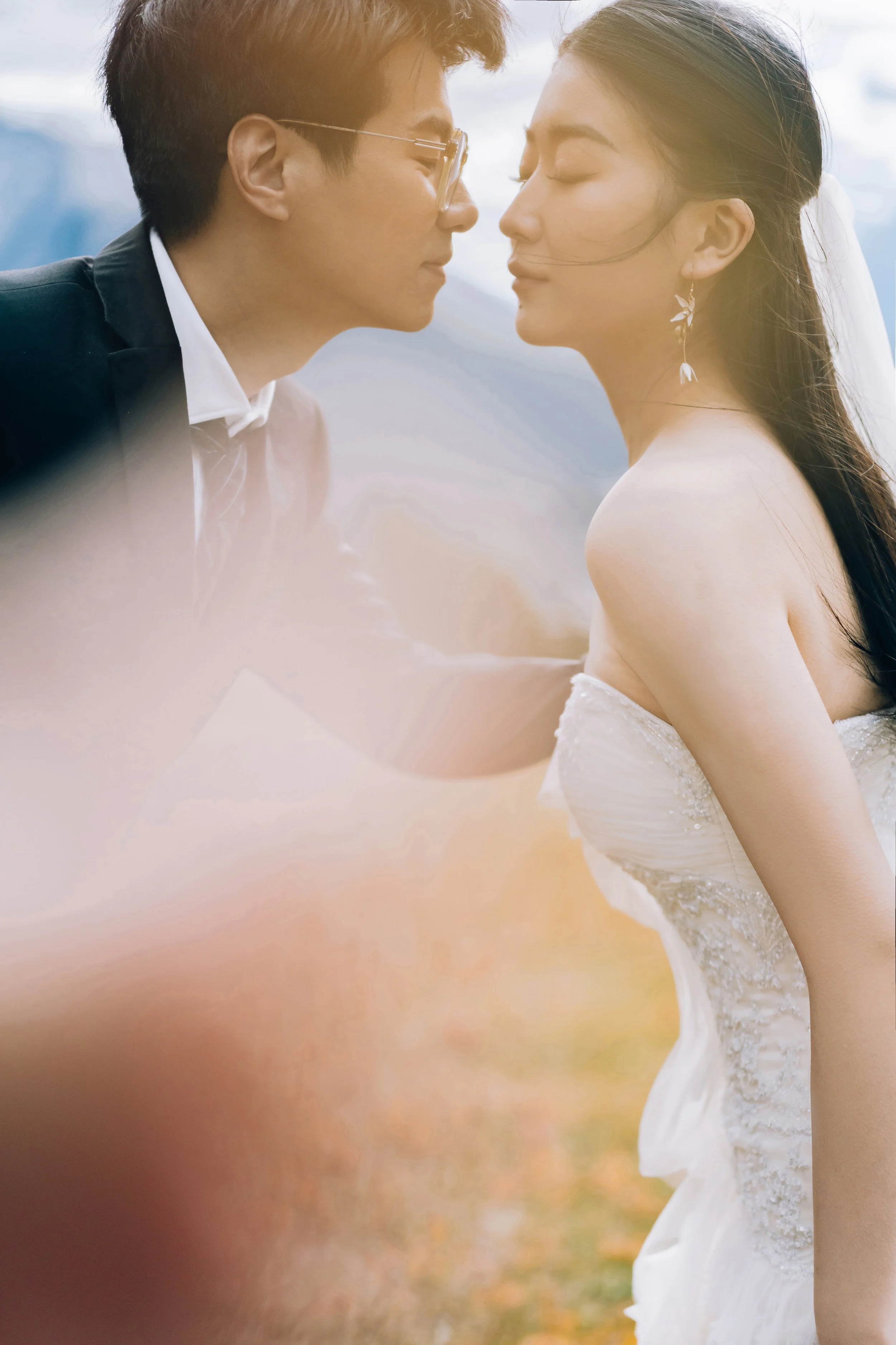 A bride and groom are about to kiss outdoors on their wedding day, the groom wearing glasses and a dark suit, the bride in a strapless white wedding gown with earrings, both with eyes closed.
