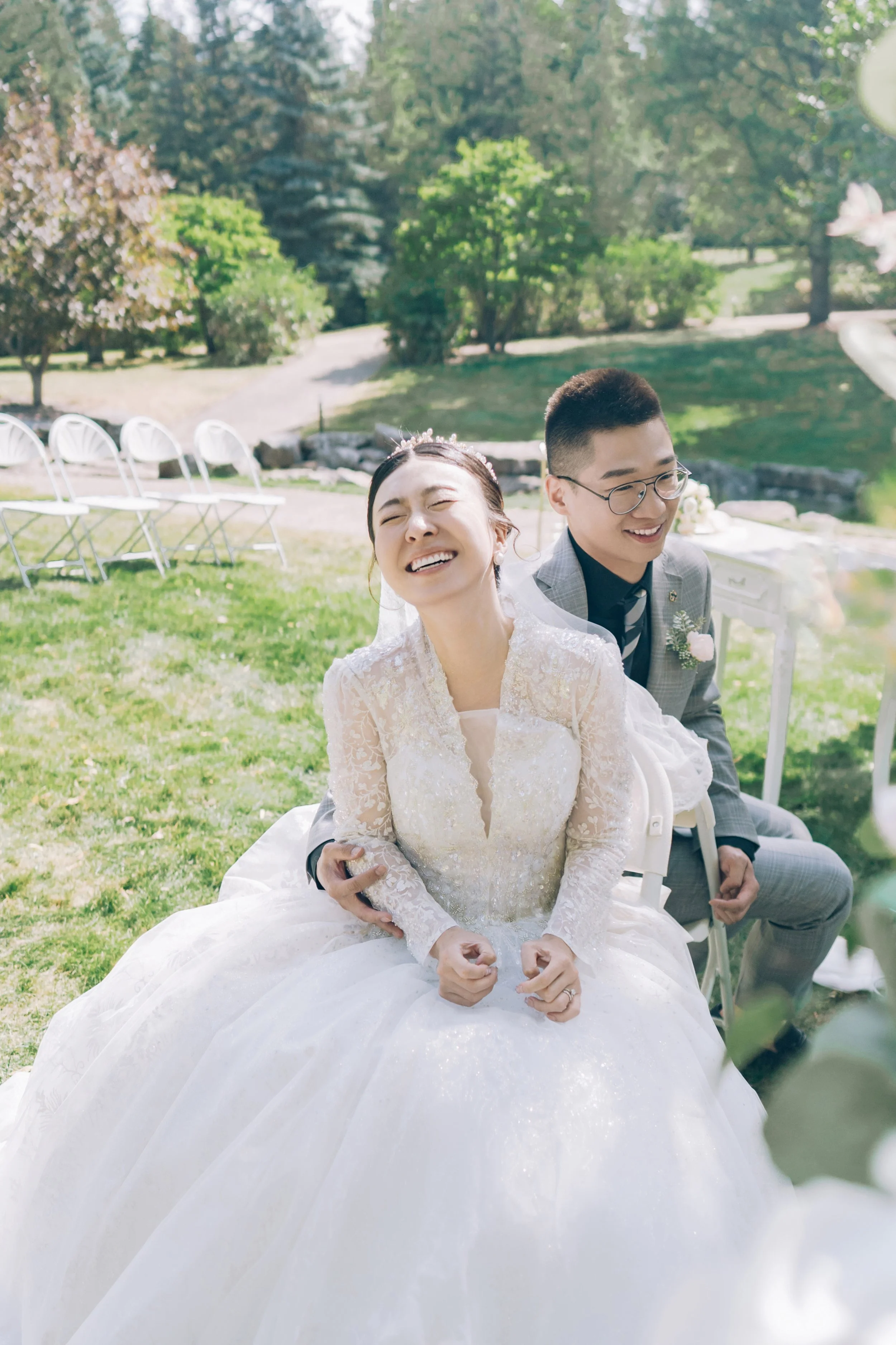 Bride and groom sitting outdoors at a wedding, smiling and laughing in a lush green garden setting.