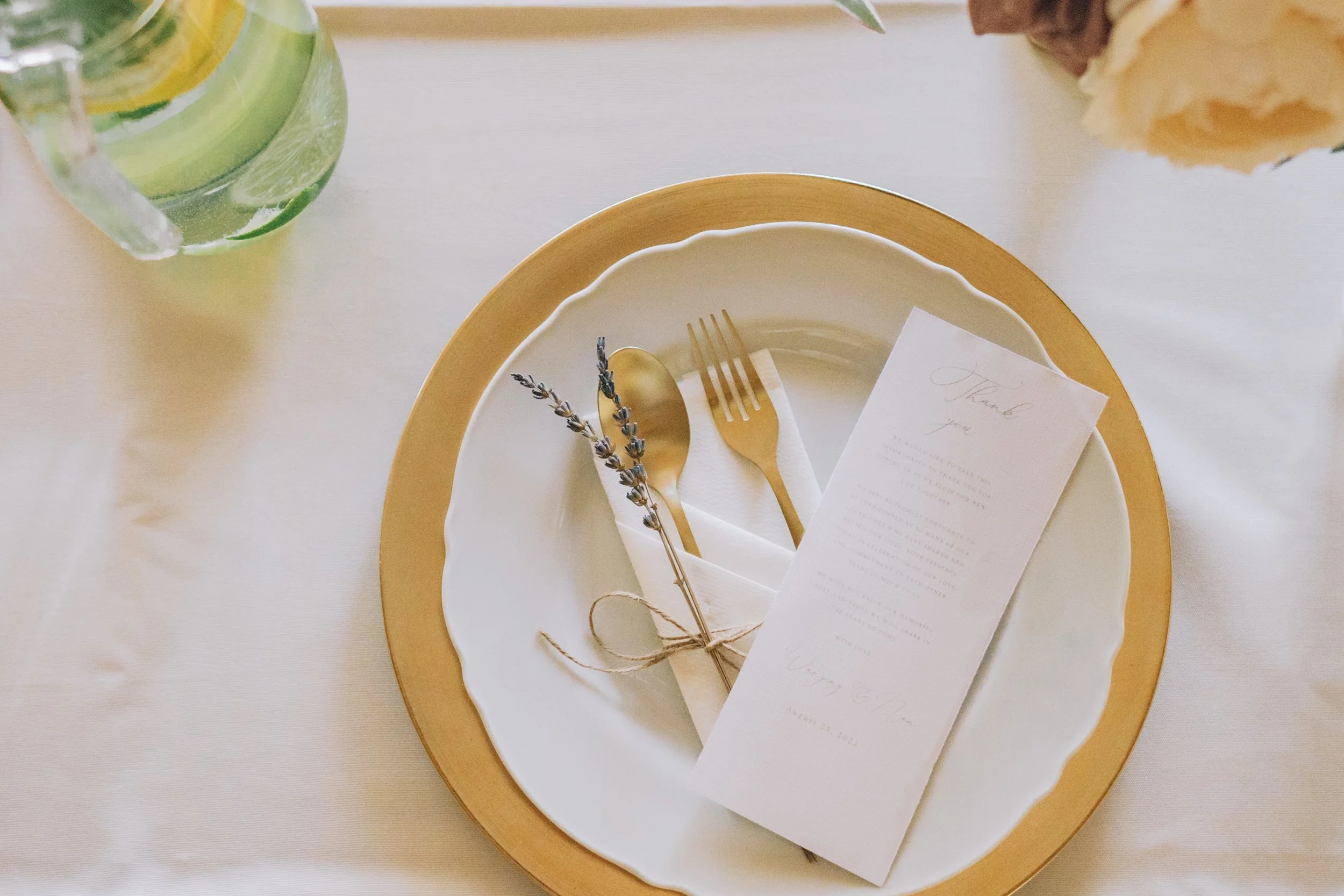 Elegant table setting with a white plate on top of a gold charger, gold fork and spoon wrapped in a white napkin with a sprig of lavender, a white thank you card, and a glass of water with lemon slices on a white tablecloth.