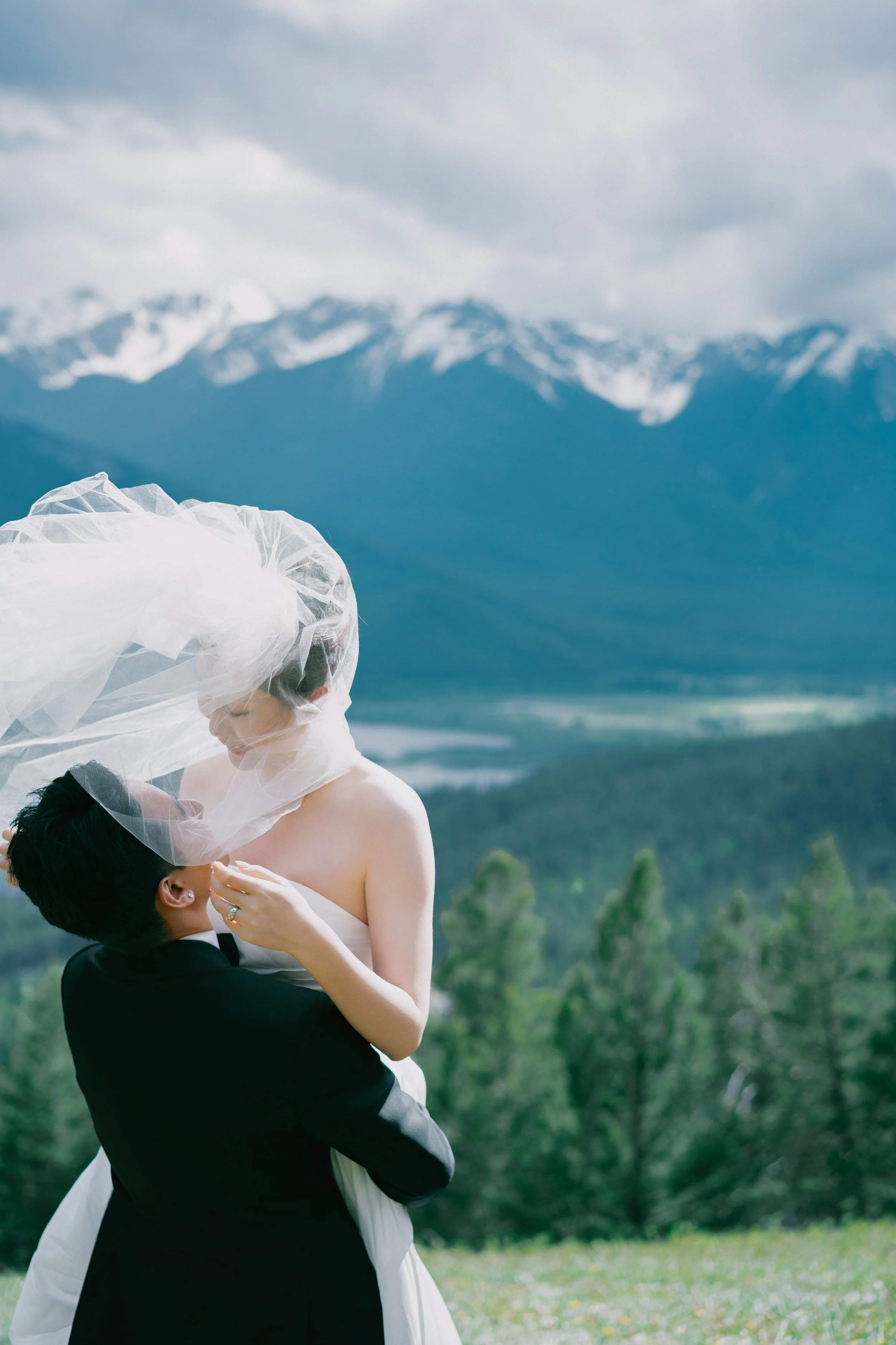 A bride and groom embrace outdoors with a mountainous landscape background, the bride's veil flowing in the wind.