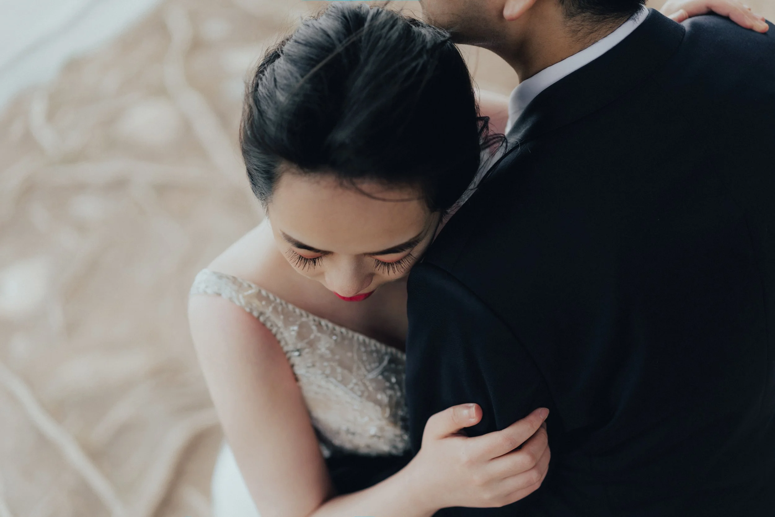 A bride and groom embrace closely, with the bride resting her head on the groom's shoulder, on a sandy surface.
