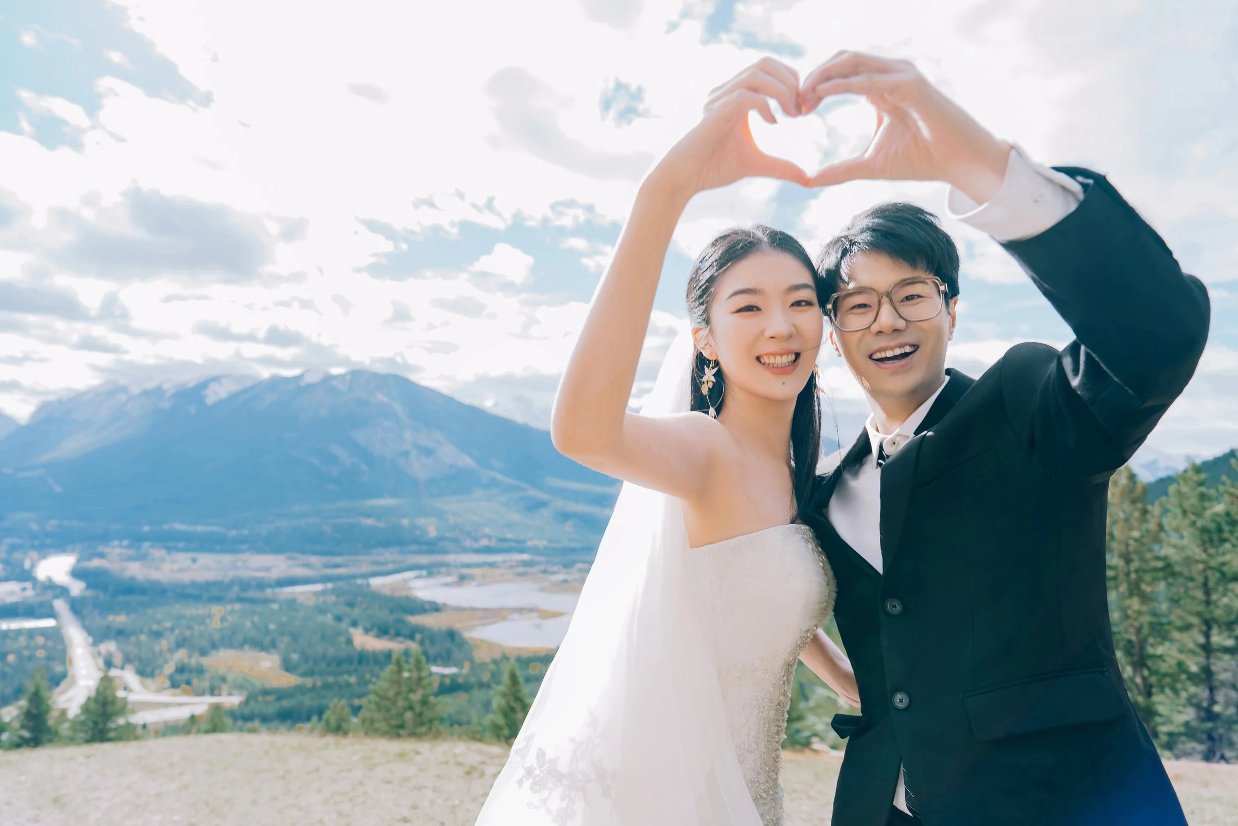 A happy bride and groom taking a selfie outdoors with a mountain and sky background, forming a heart shape with their hands.