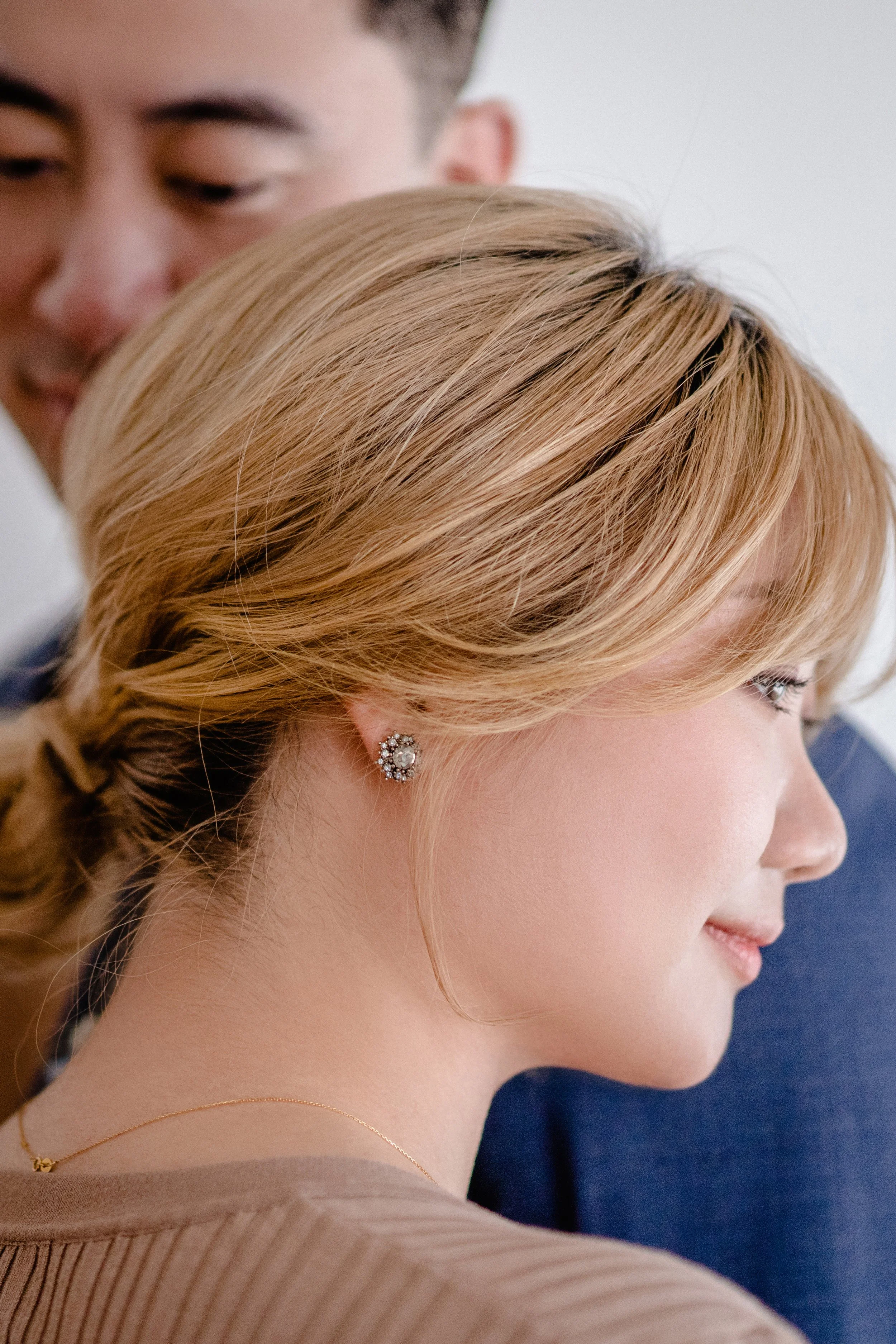 Close-up of a woman with blonde hair in an elegant updo, wearing pearl and diamond earrings and a thin gold necklace, with a man in the background.