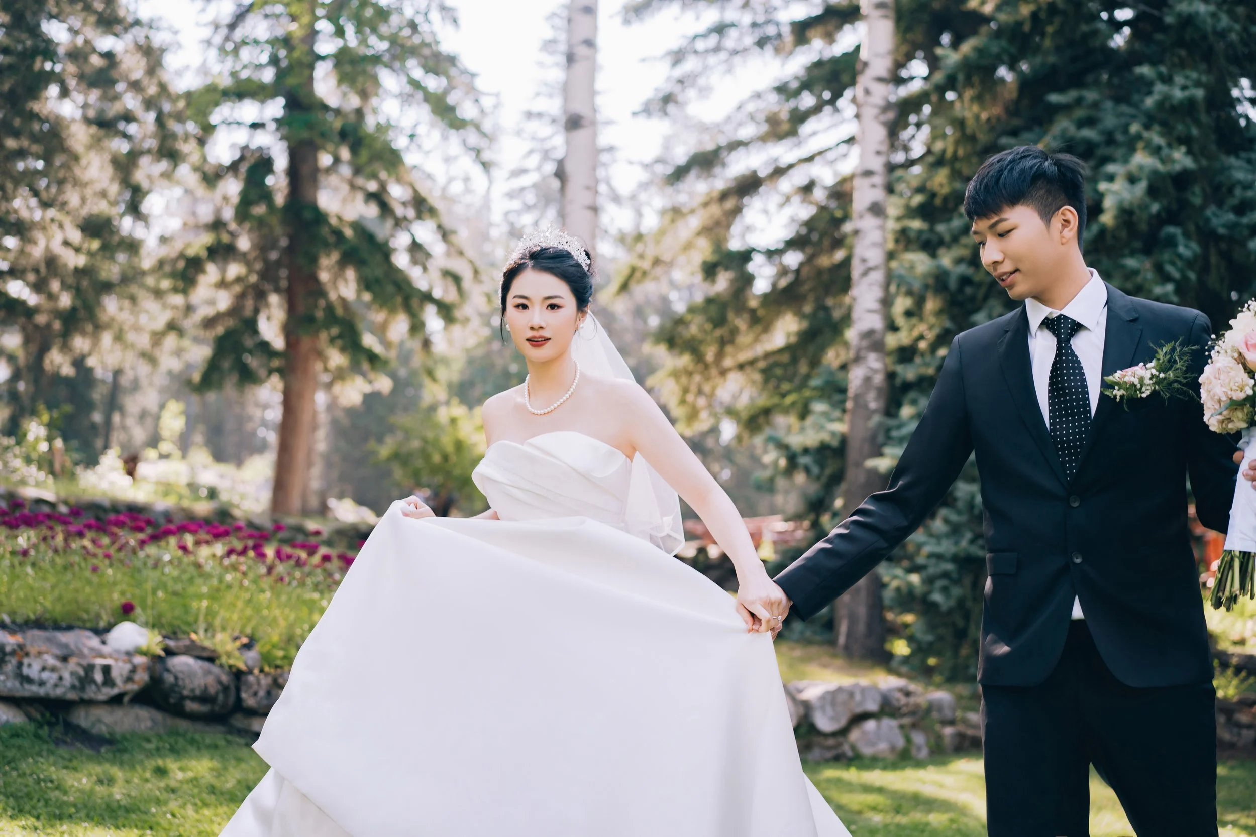 A bride and groom holding hands in a park during their wedding, with trees and flowers in the background.