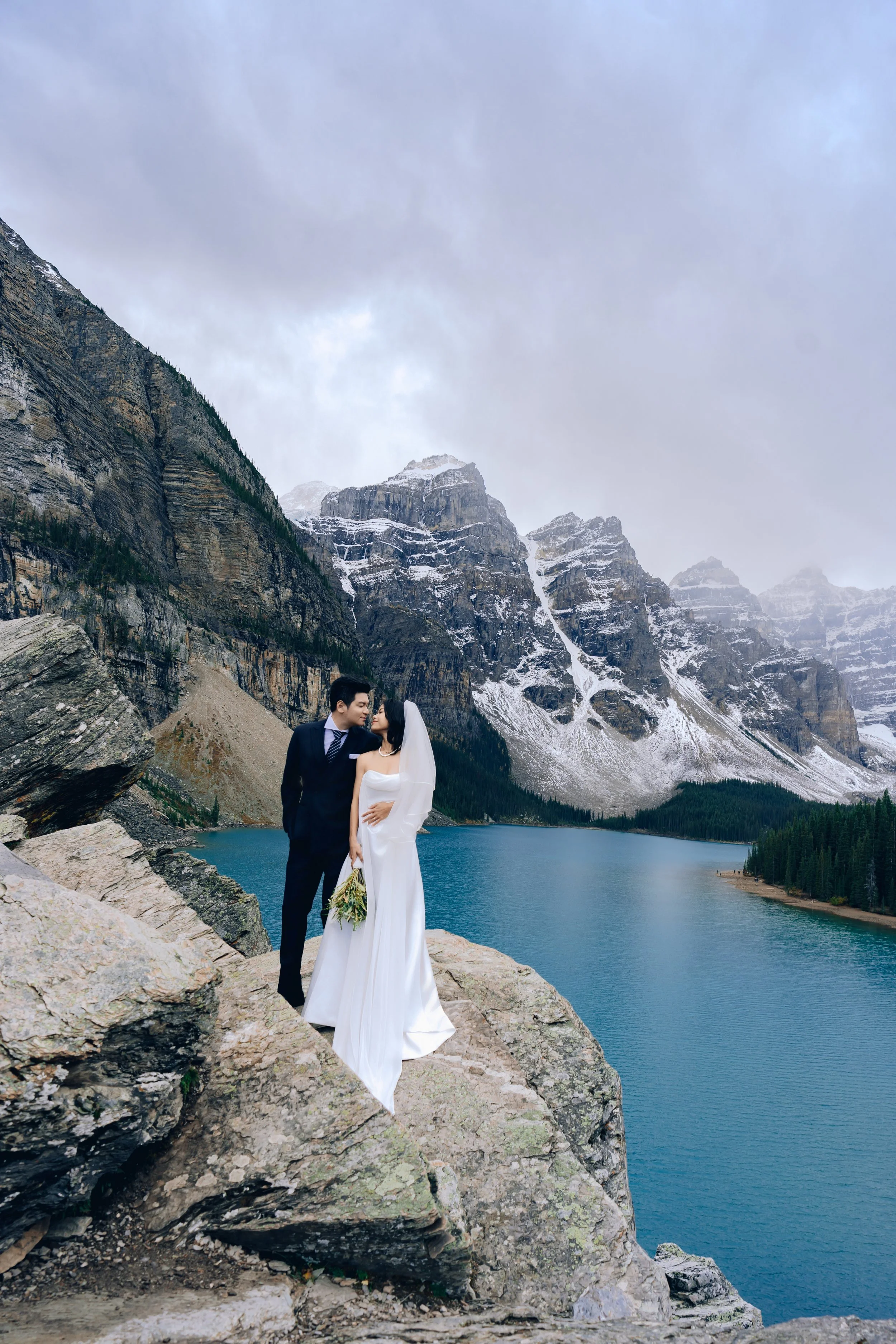 A bride and groom in wedding attire stand on rocks beside a lake, with mountains and cloudy sky in the background.