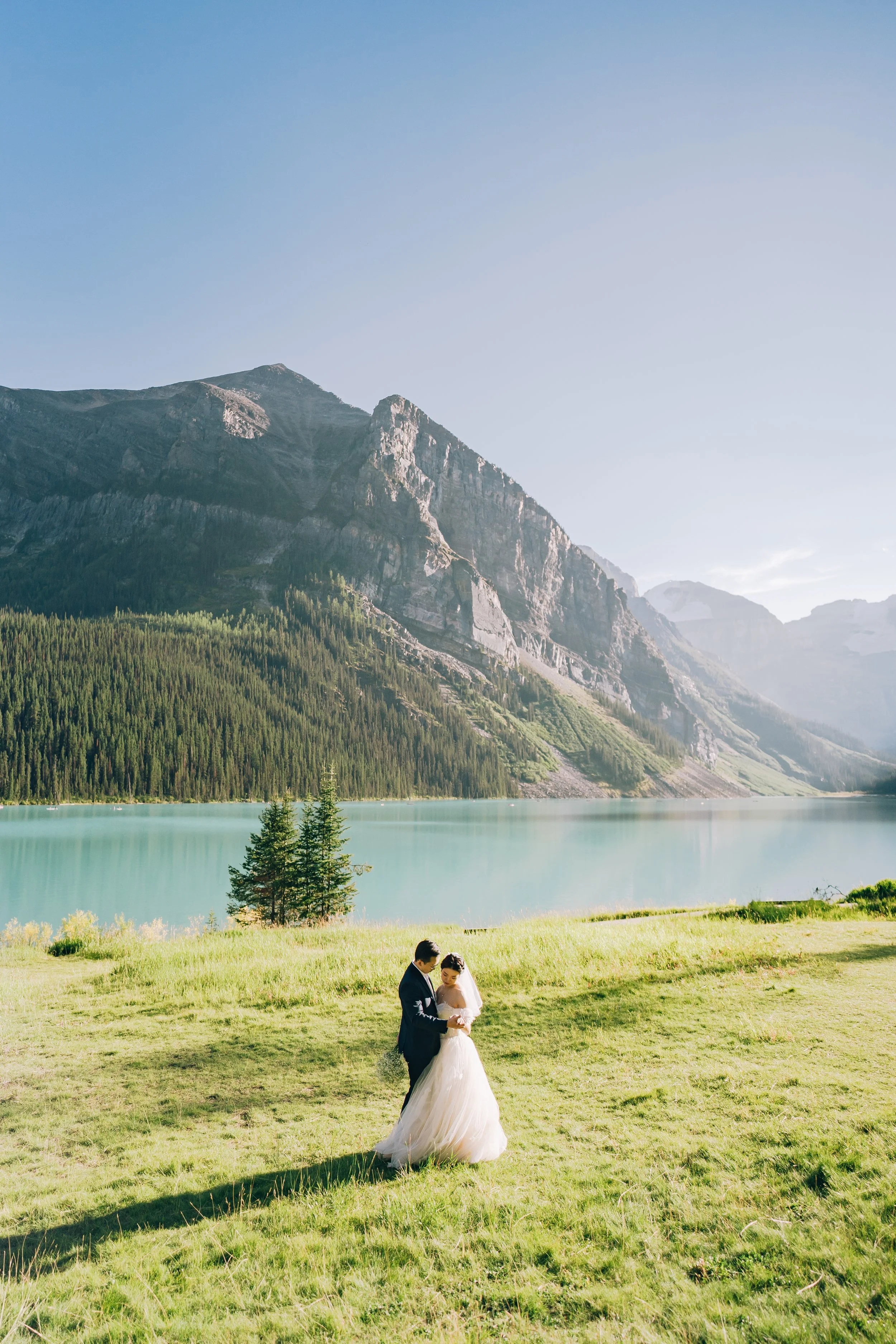 A bride and groom standing on green grass near a lake with mountains in the background