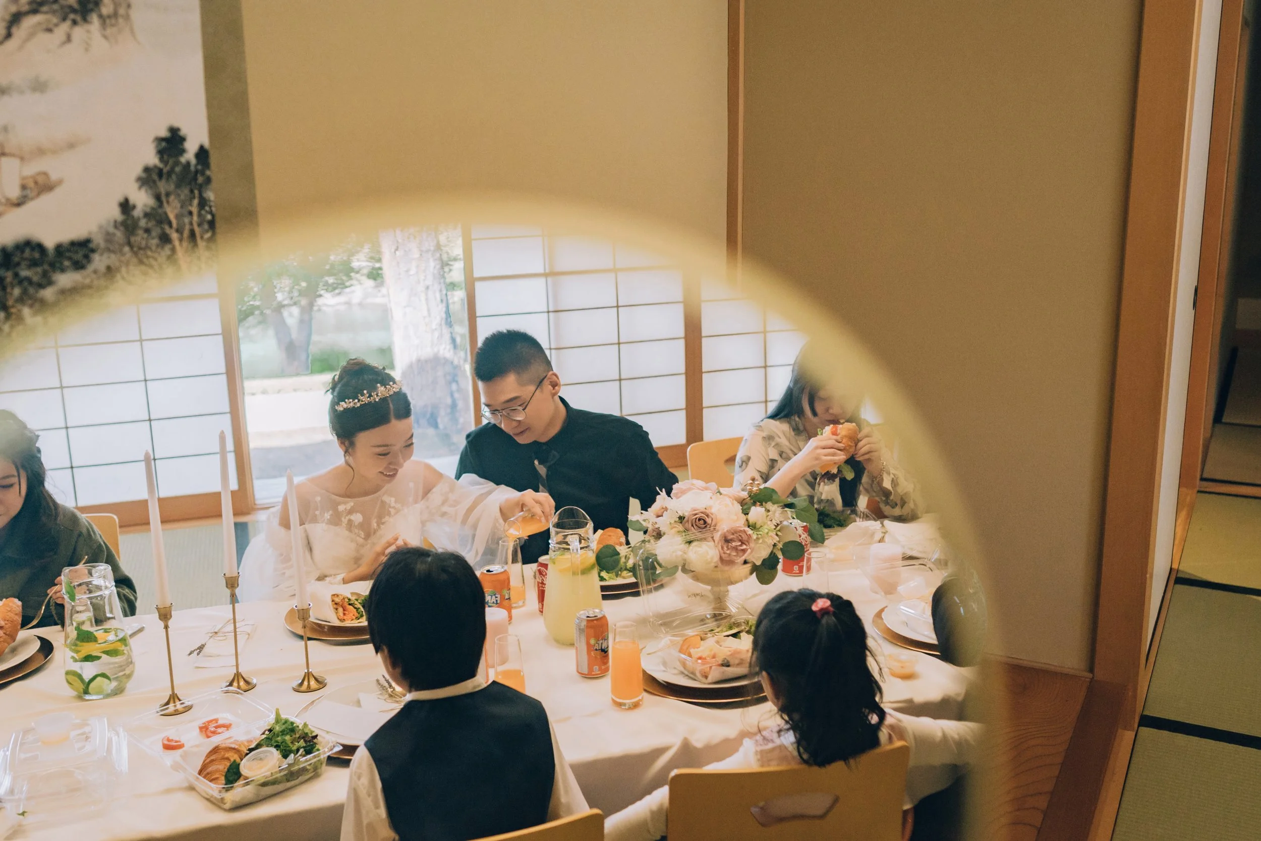 People celebrating at a dining table viewed through a round cutout in a wall, with a bride in a white wedding dress, a man in black, children, and various dishes and drinks on the table.