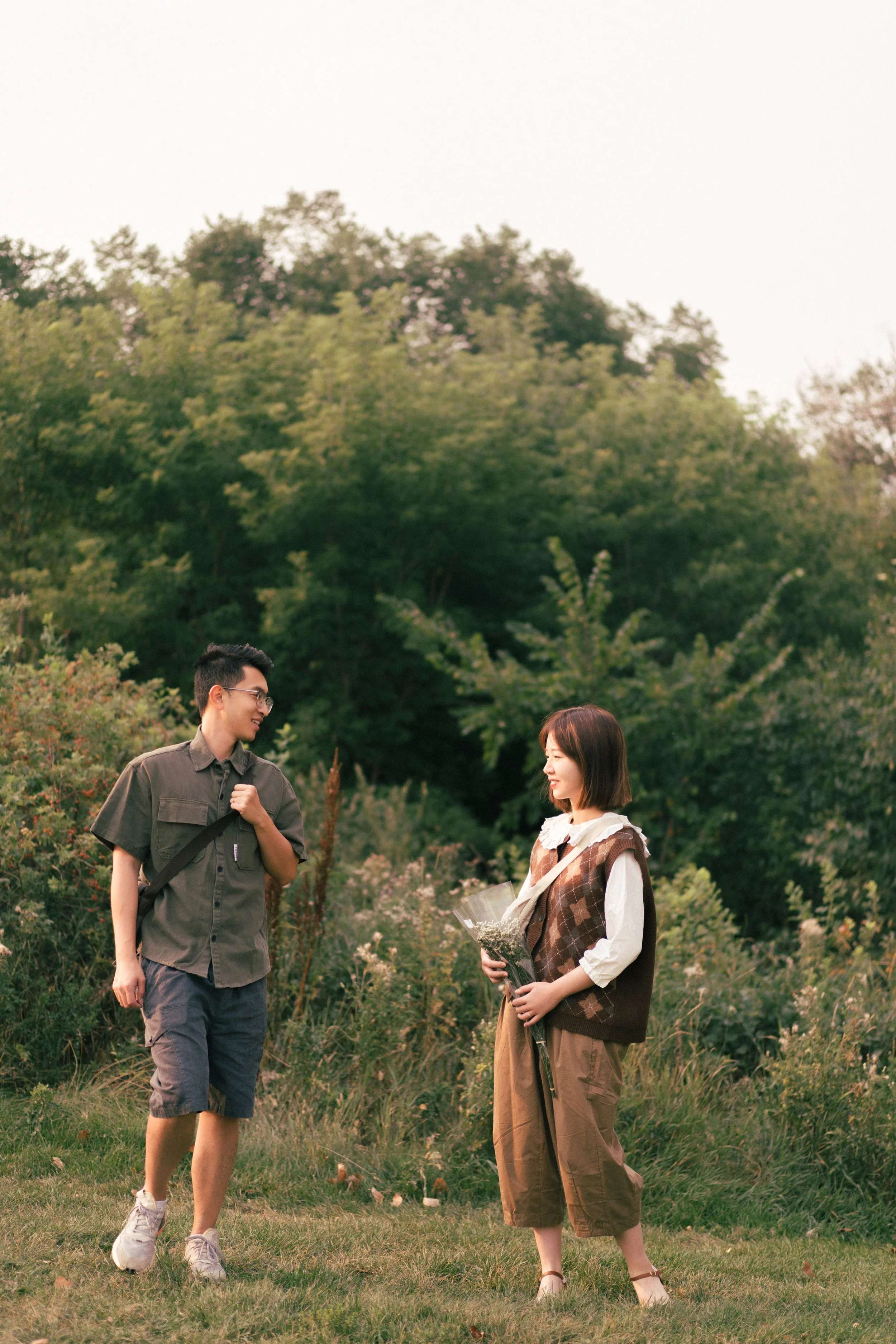 A young man and woman exchanging flowers outdoors in a grassy area with trees in the background.