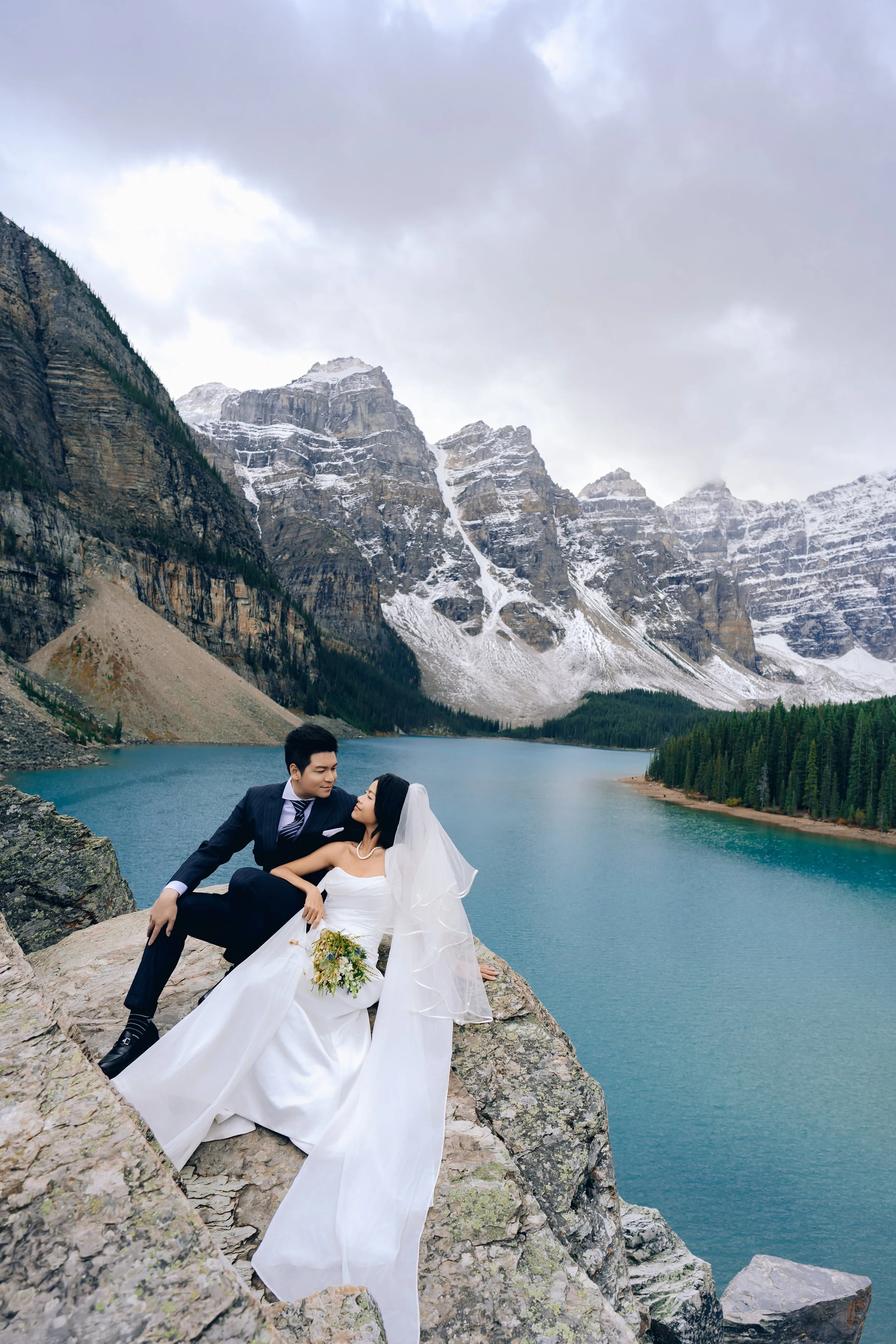 A bride and groom in wedding attire sitting on rocks by a lake surrounded by mountains and pine trees.
