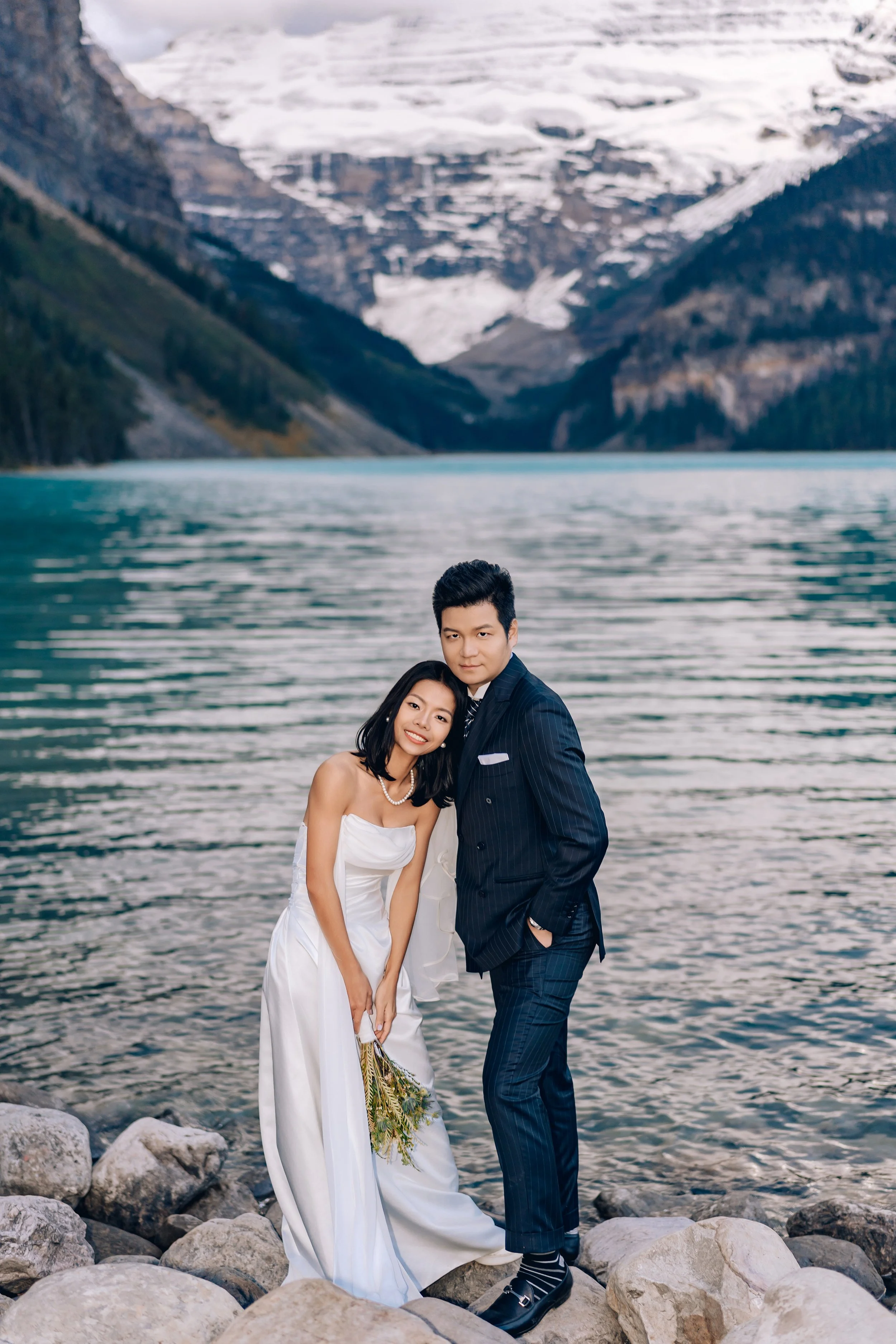 A couple dressed in wedding attire standing on rocks by a lake with mountains in the background.