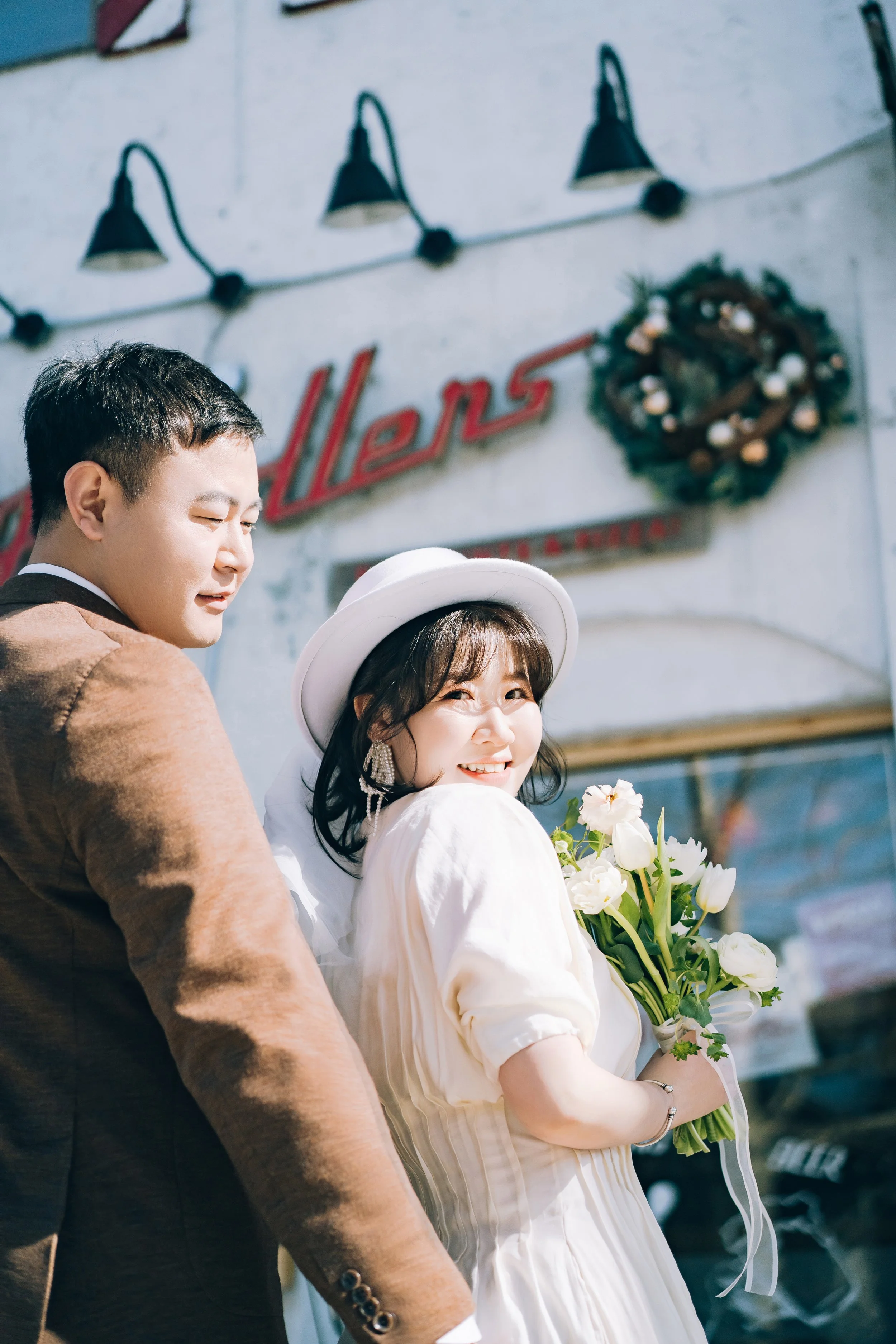 A smiling woman holding a bouquet of white flowers, wearing a white hat and a white dress, standing next to a man in a brown jacket, outside a building with a sign that says 'Hellers' and a holiday wreath, on a sunny day.