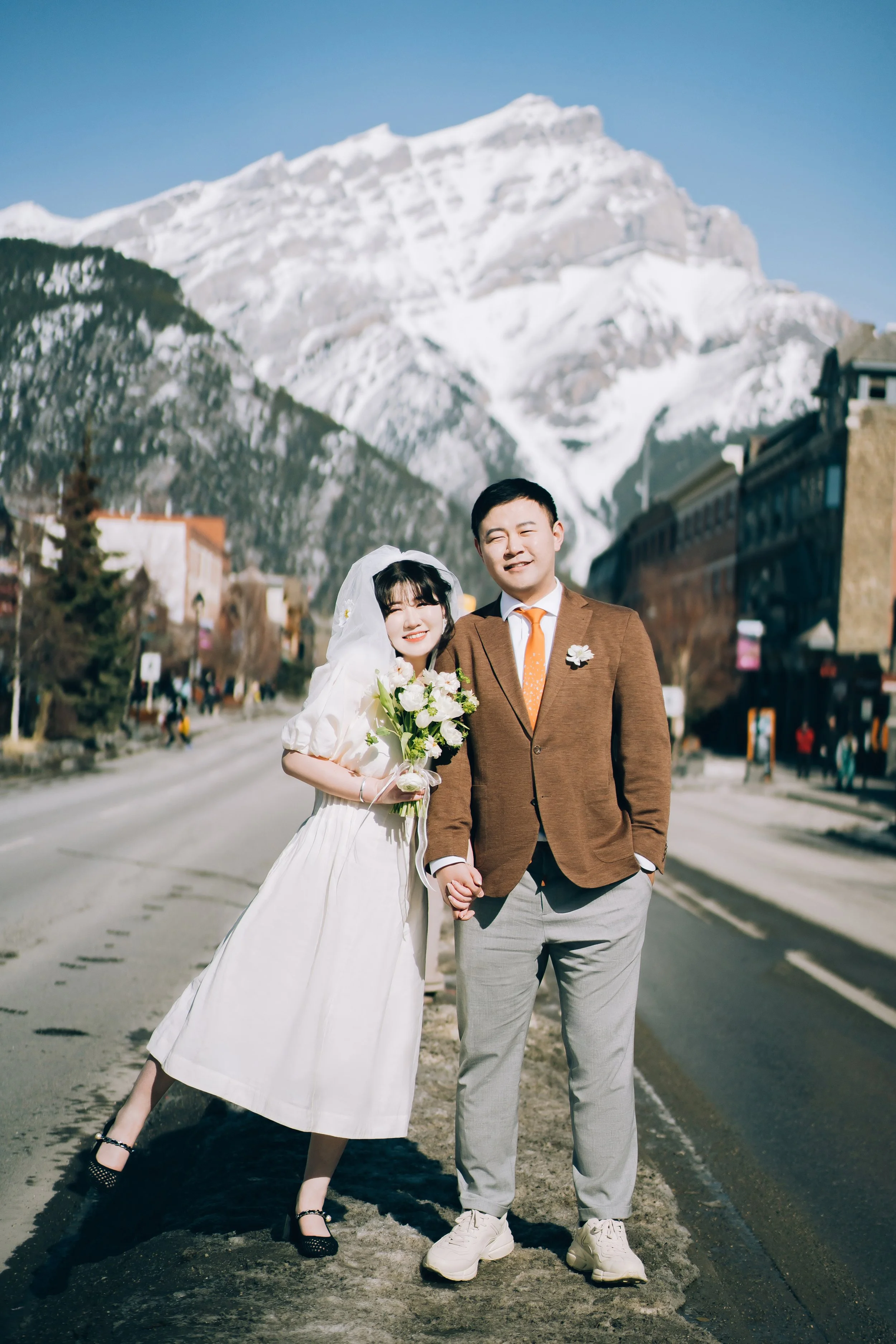 A happy bride and groom holding hands and smiling, standing on a snow-capped mountain street on their wedding day, with mountains and buildings in the background.