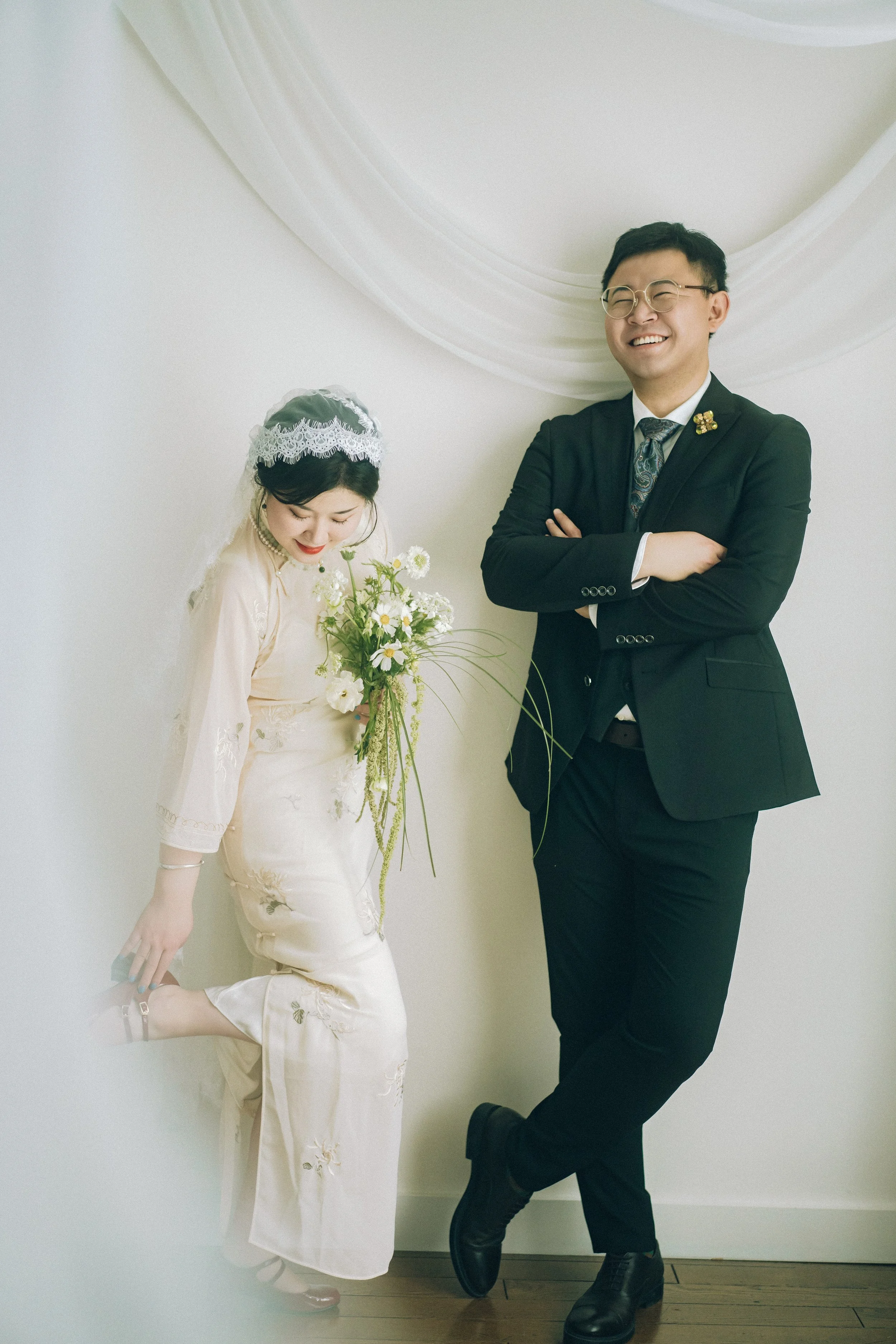 A bride in a cream-colored traditional dress holding a bouquet of white and green flowers, and a groom in a black suit with glasses standing against a white wall with draped fabric, both smiling.