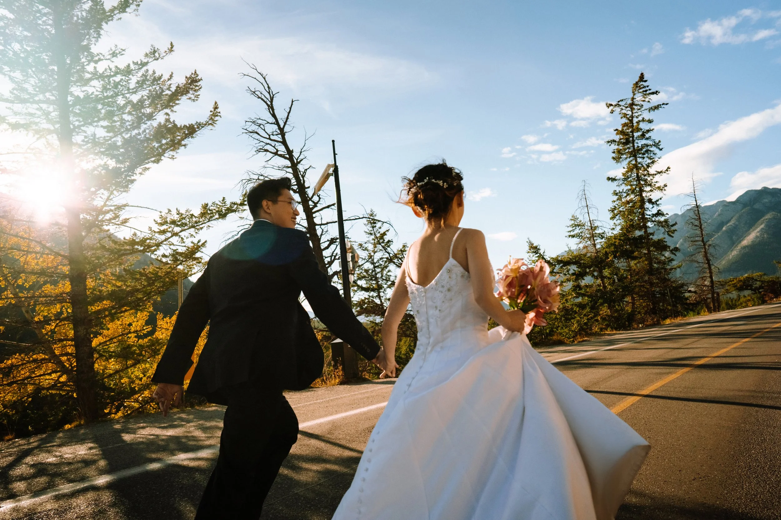 Bride and groom holding hands, running on a scenic road, surrounded by trees and mountains, with the sun shining from behind.