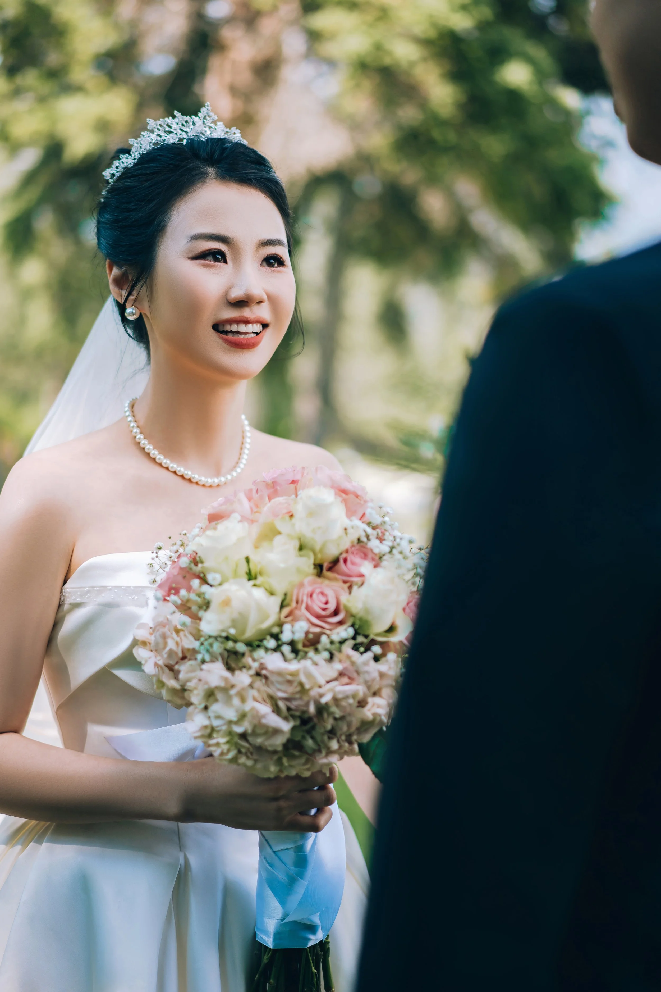 A bride in a strapless white wedding gown holding a bouquet of pink and white roses, smiling at her groom outdoors with trees in the background.