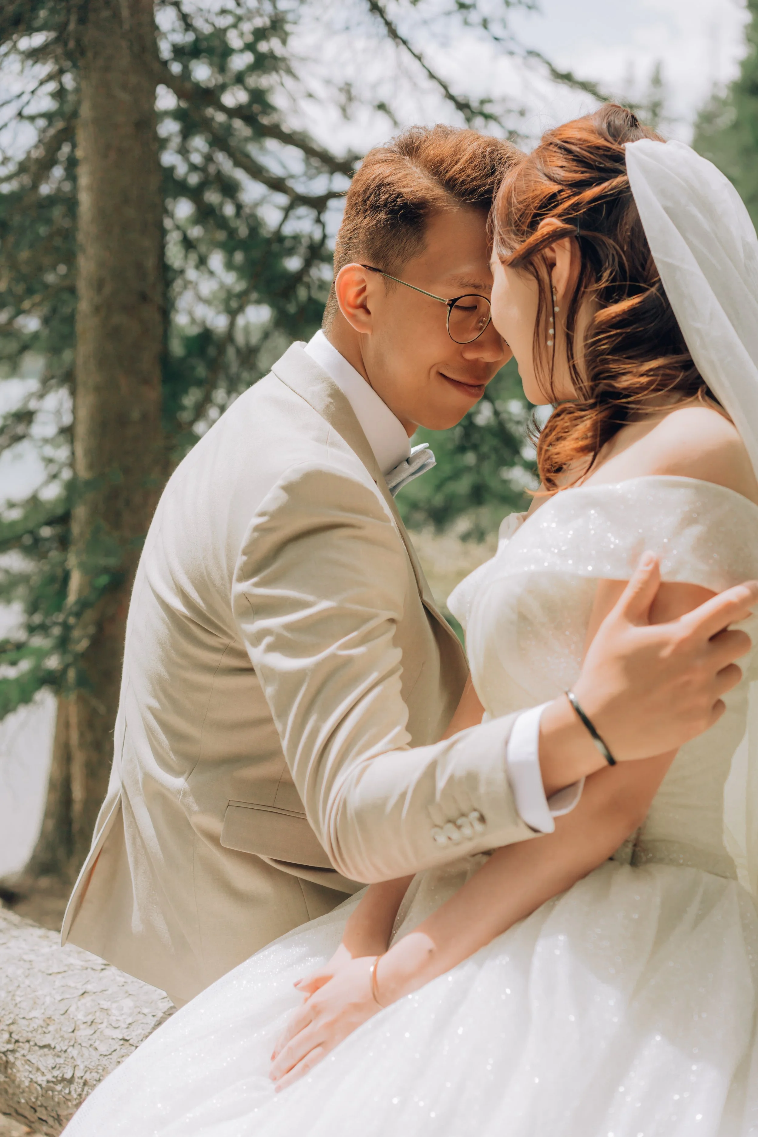 A couple in wedding attire share a close, intimate moment outdoors with trees in the background.