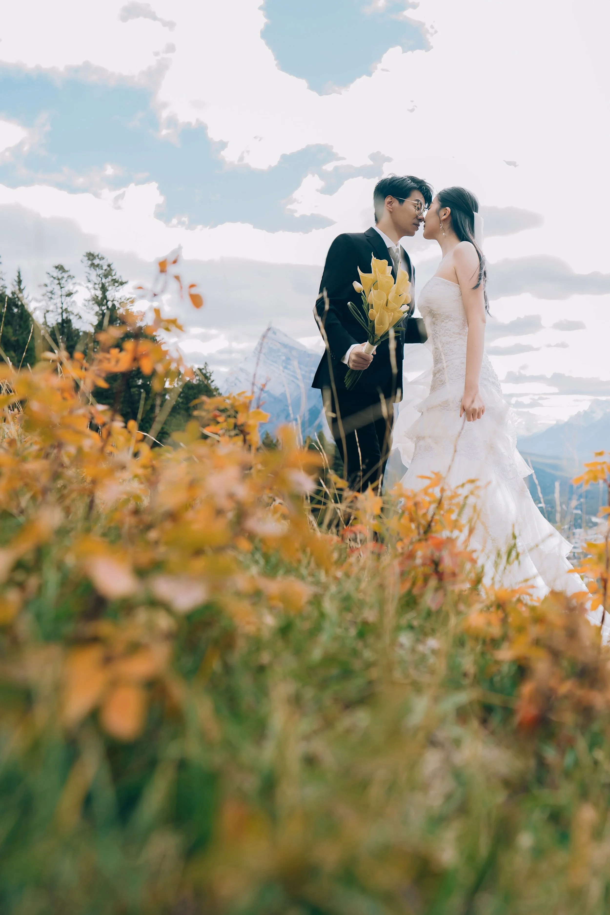 A bride and groom sharing a moment outdoors with mountains in the background, the groom is holding a bouquet, and they are standing among orange and green foliage.