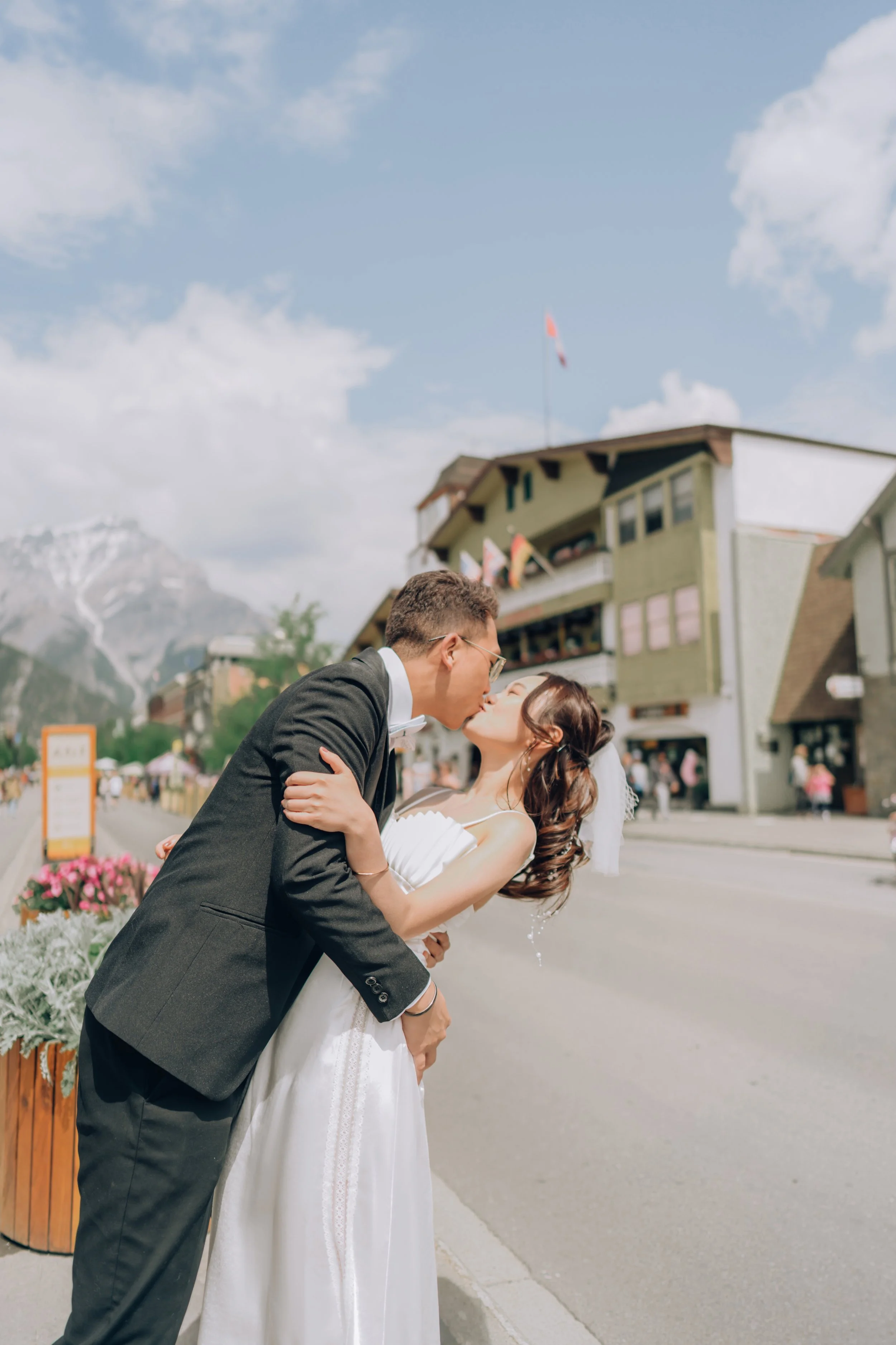 A newlywed couple in wedding attire sharing a kiss on a street with mountains in the background.