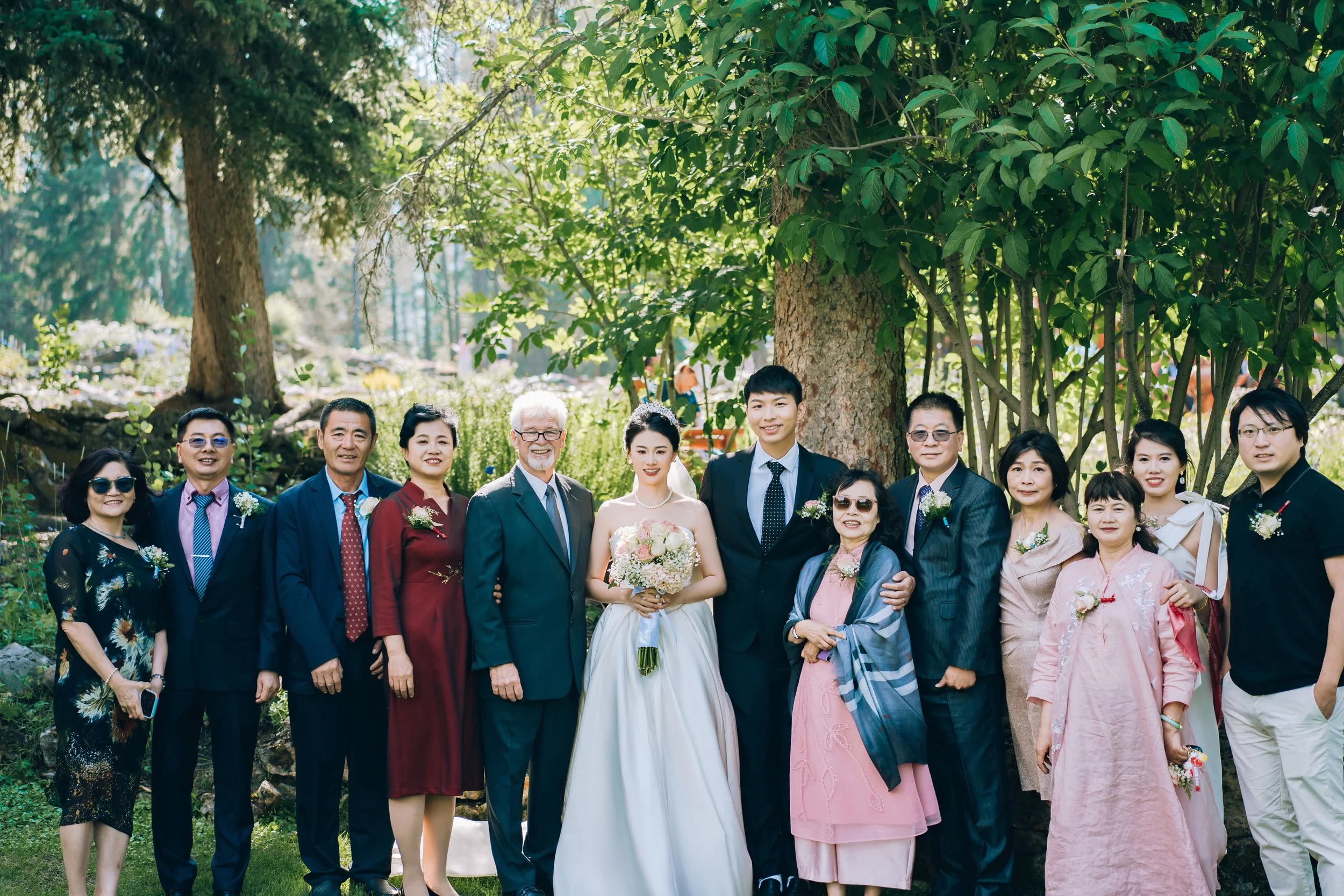 Group of wedding guests and couple standing outdoors in front of a large tree, smiling, dressed in formal and traditional attire.