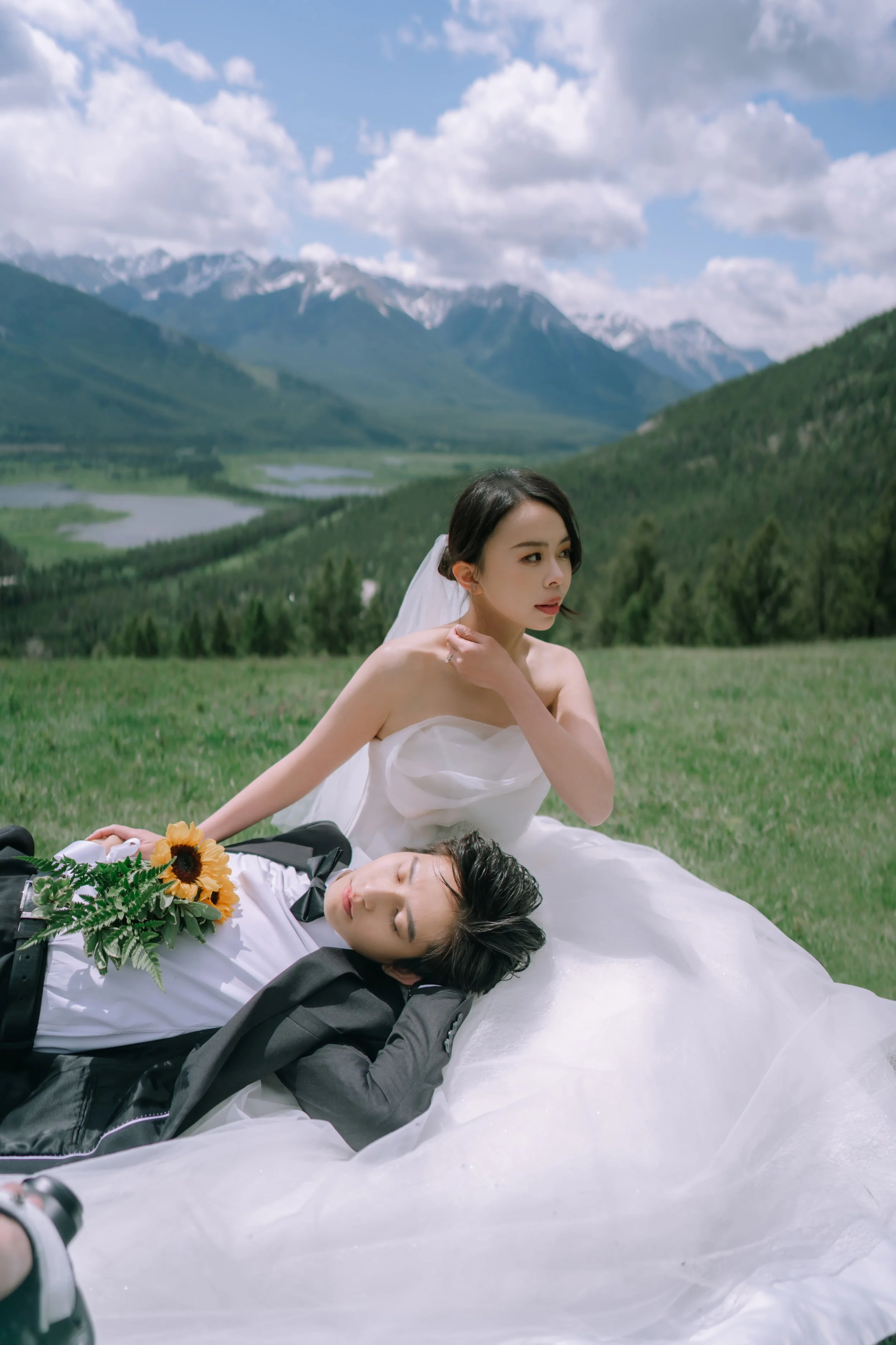 A woman in a wedding dress and a man in a suit lying on a grassy field in the mountains, with the woman sitting up and the man resting his head on her lap, holding a bouquet of sunflowers.