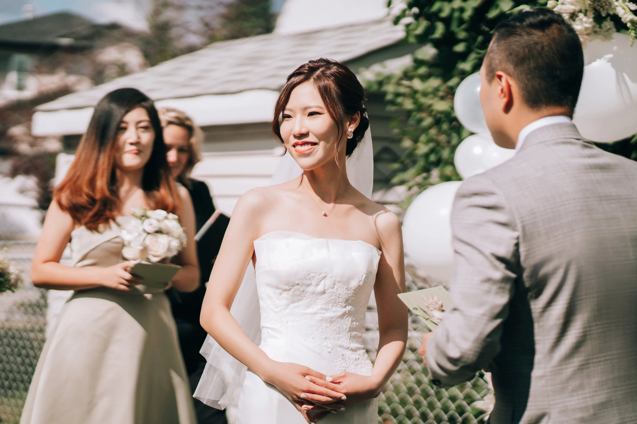 A bride in a strapless white wedding gown smiling at her groom during her wedding ceremony outdoors, with bridesmaids holding bouquets in the background.