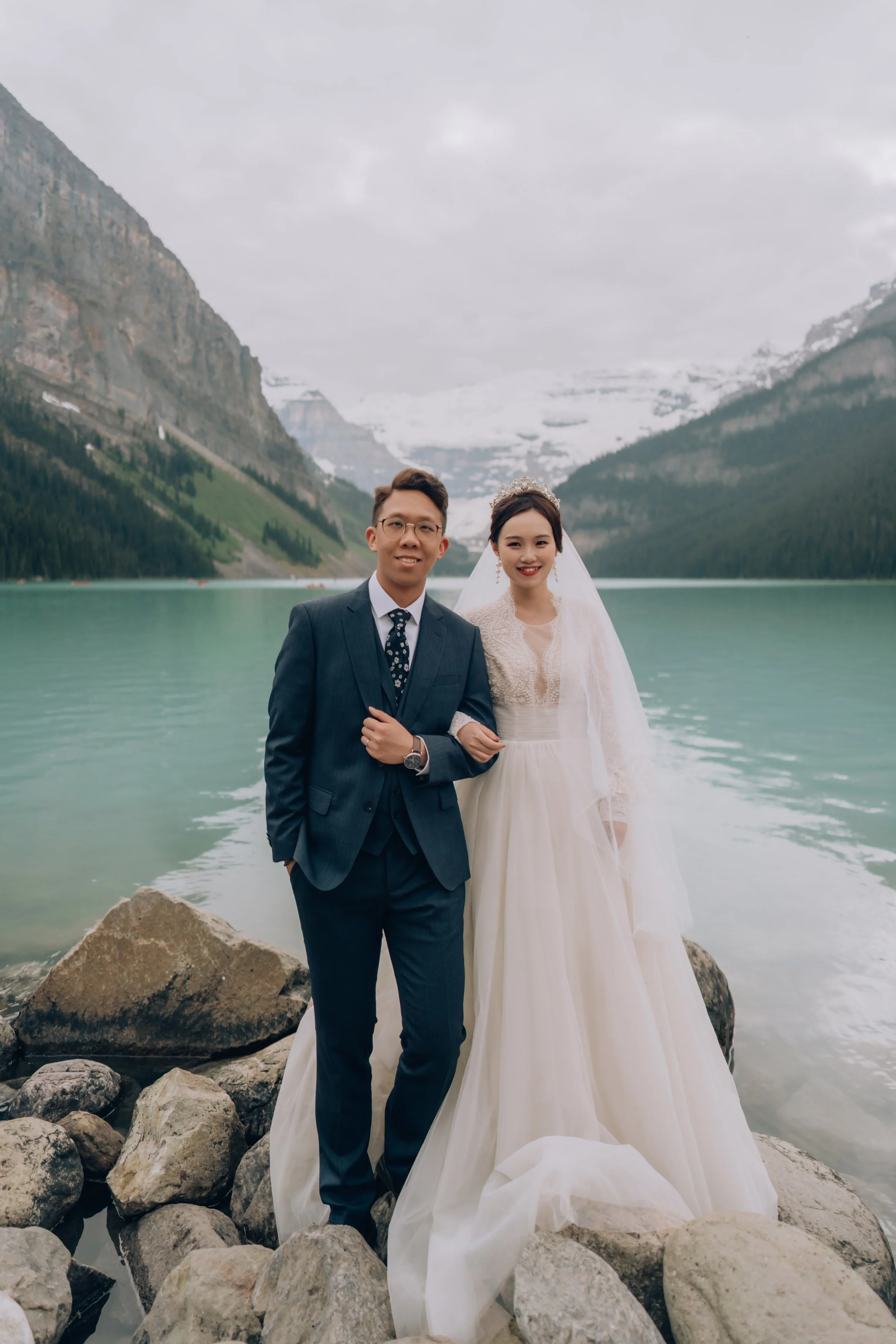 A bride and groom in wedding attire standing on rocks by a lake at the base of snow-capped mountains, smiling at the camera.
