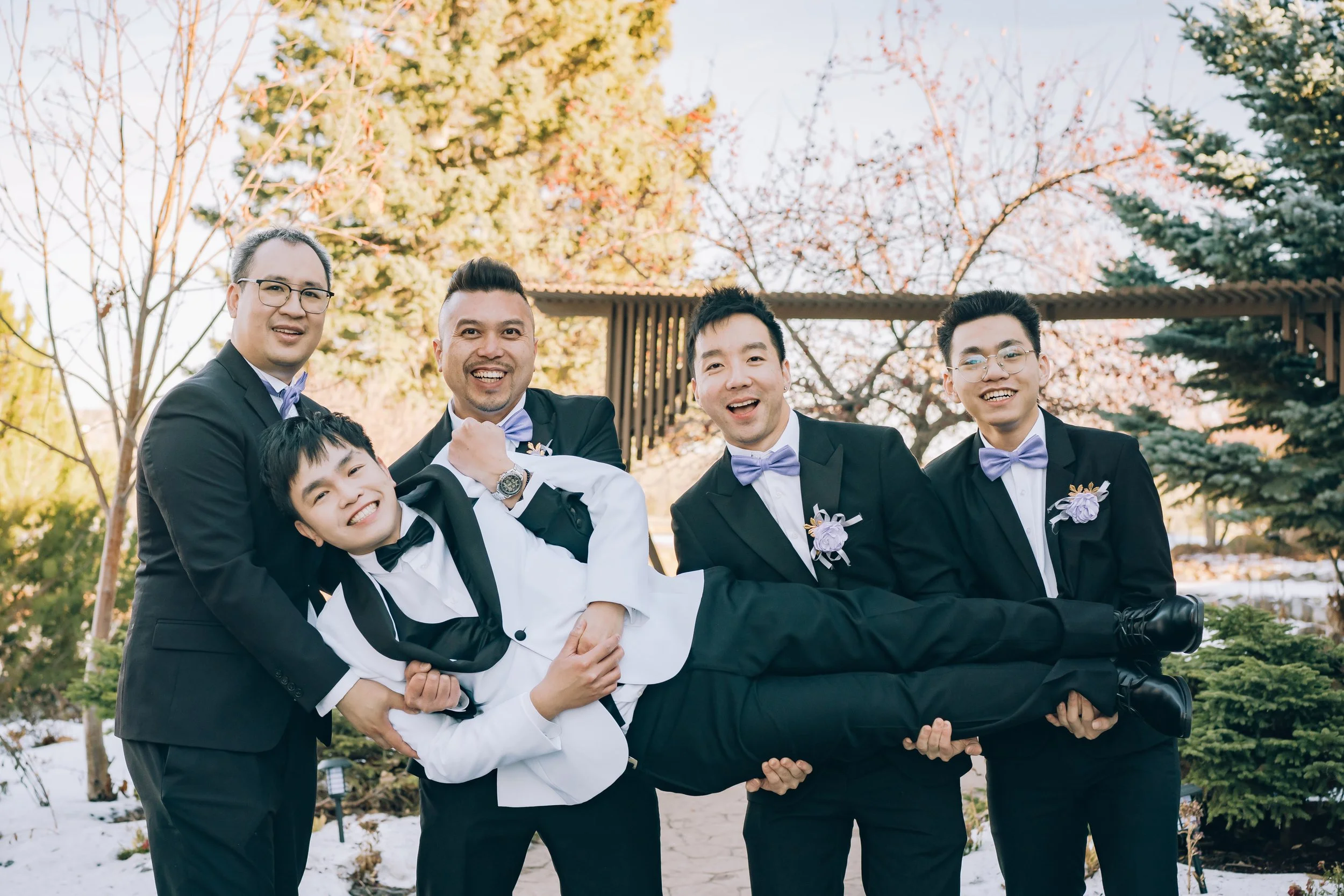 Group of five men in suits and bow ties, smiling and holding a young boy in a tuxedo, outdoors near trees and snow.