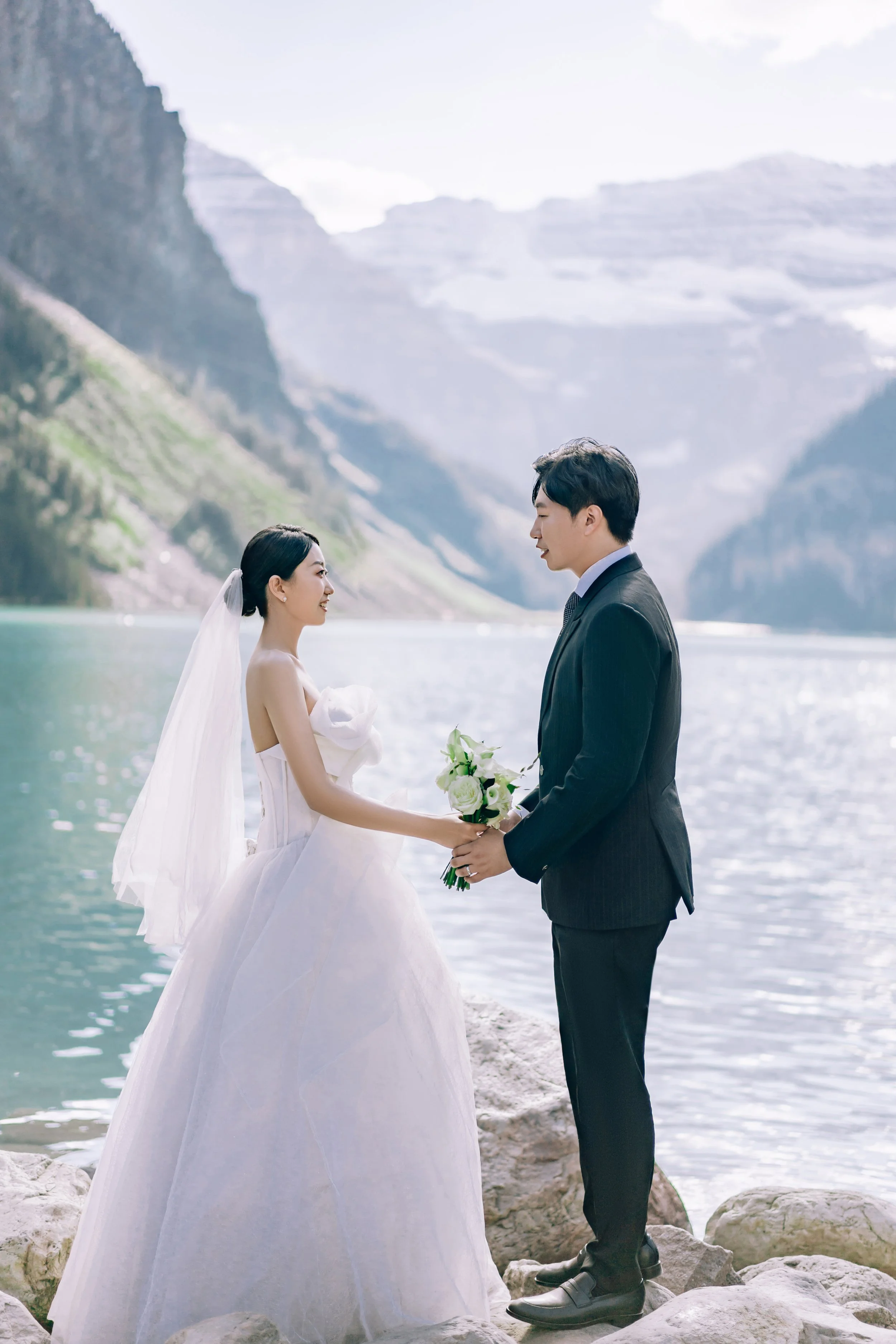 A bride and groom standing on rocks by a lake with mountains in the background, holding a bouquet and facing each other during their wedding photos.