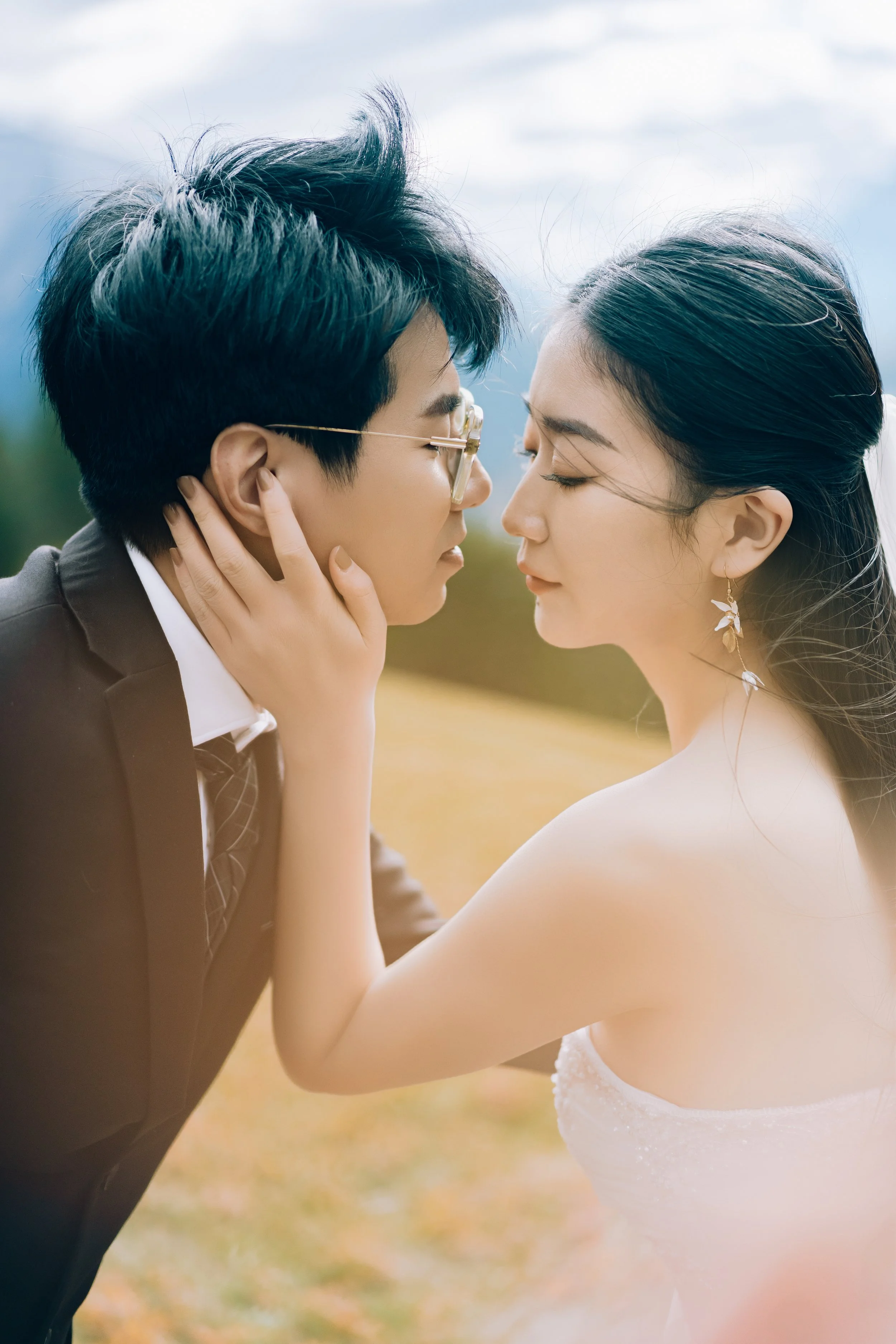 A couple with closed eyes face closely, the woman gently holding the man's face, outdoors with a blurred natural background.