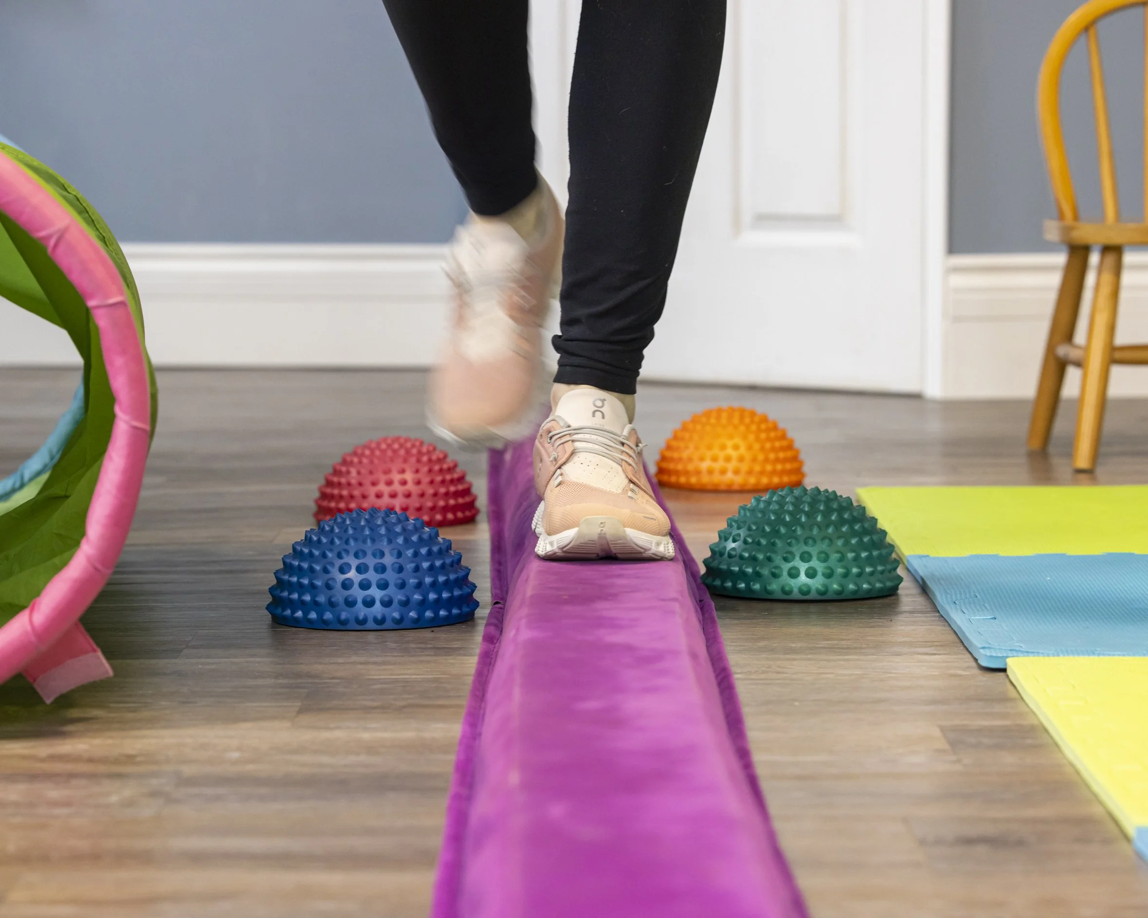 A person's legs and feet are pictured in black pants and pink shoes walking on a purple balance beam with colourful balance toys in the background.