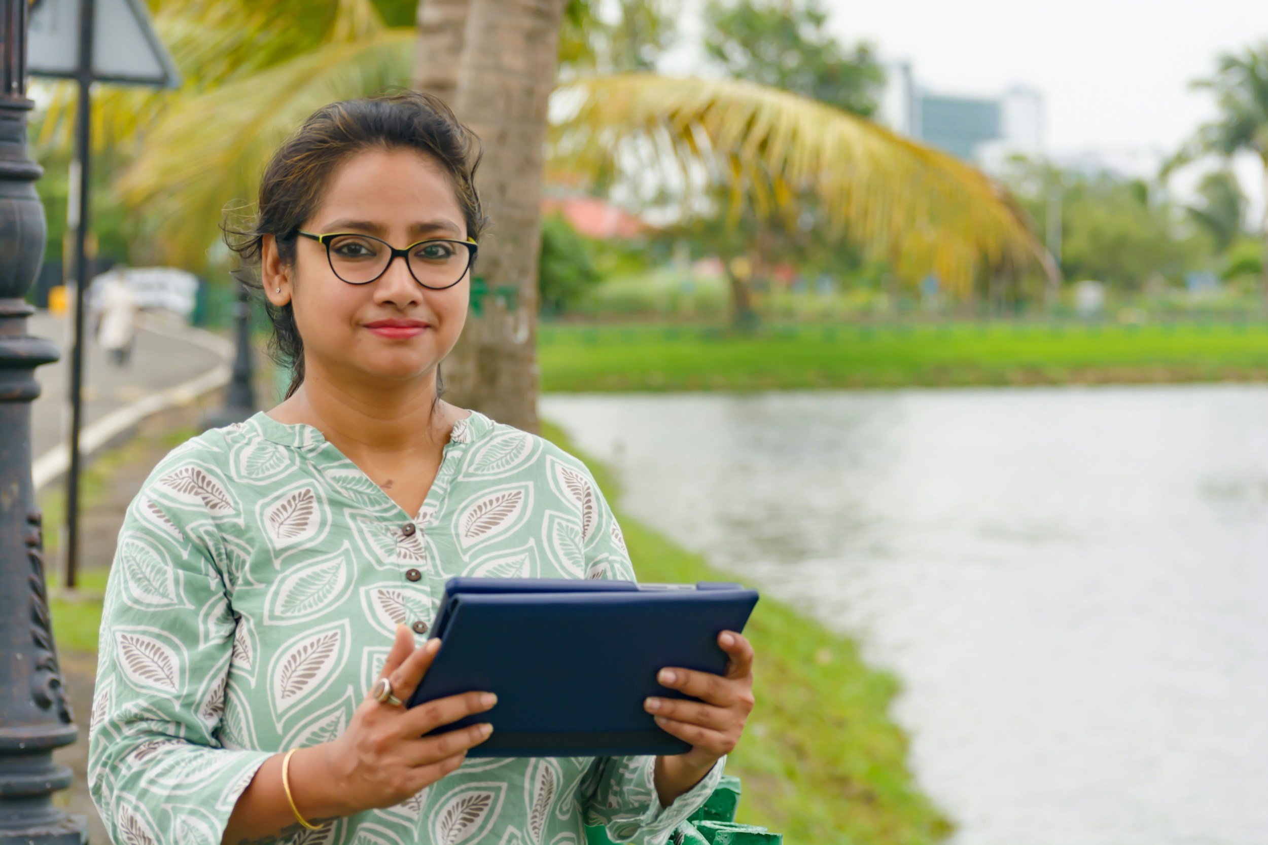 A woman with glasses and a patterned green and white top holding a tablet near a river, with trees and buildings in the background.