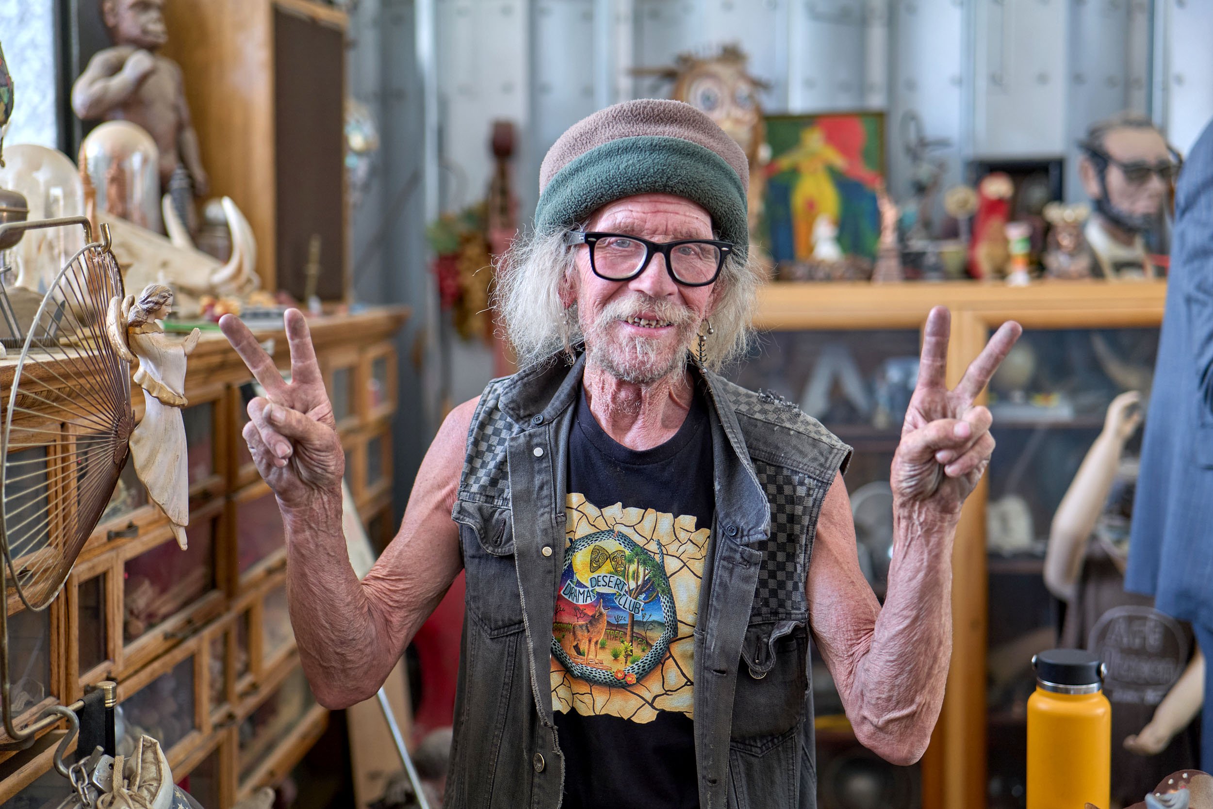 An elderly man with long gray hair, wearing glasses, a multicolored beanie, a sleeveless vest, and a black T-shirt with a desert-themed logo, making peace signs with both hands inside a shop with wooden shelves filled with various art and quirky obje