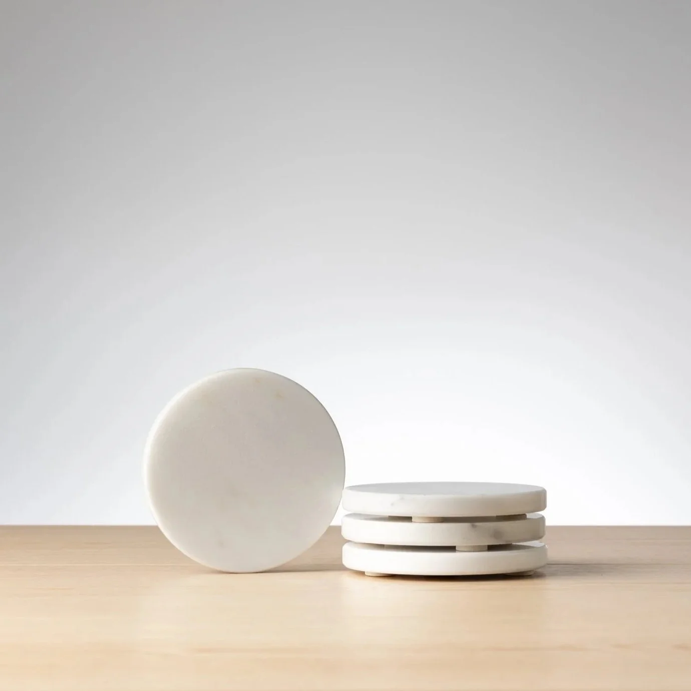 Marmo - A stack of four white marble coasters on a wooden surface with a plain white background.