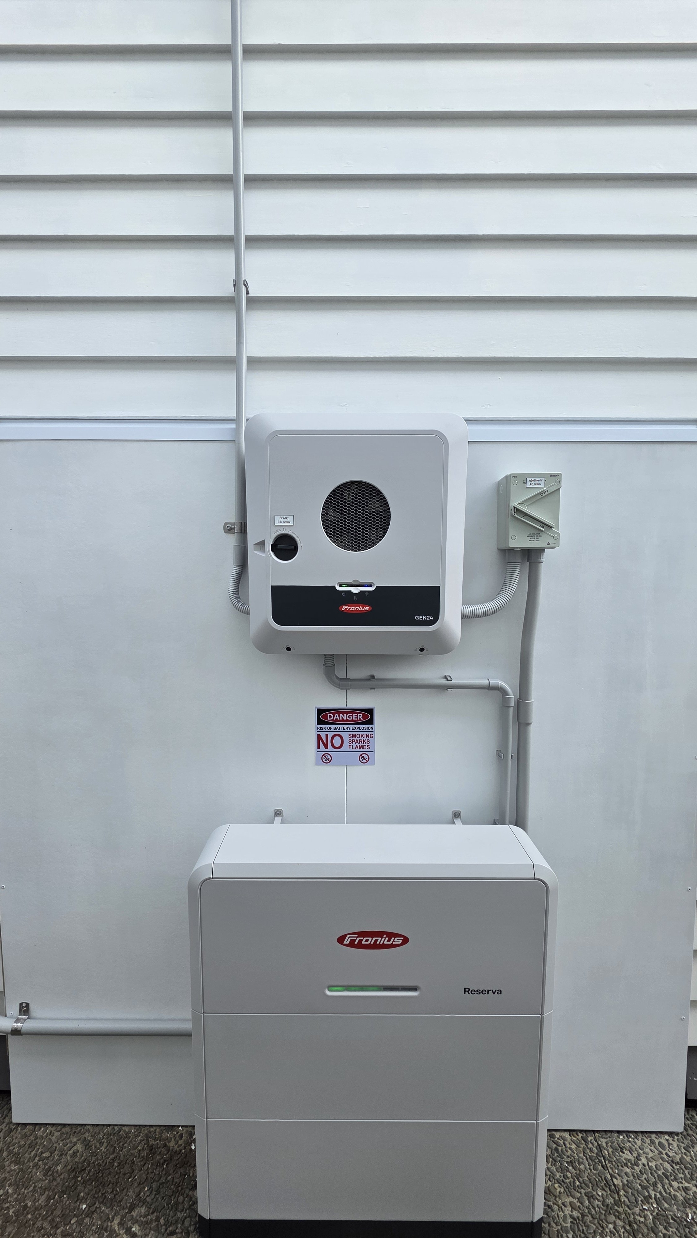 A white outdoor electrical box system with a warning sign, mounted on a white wall with horizontal siding. The system includes a larger rectangular box with a small display, a smaller box with a switch, and an insulated conduit running vertically and horizontally.