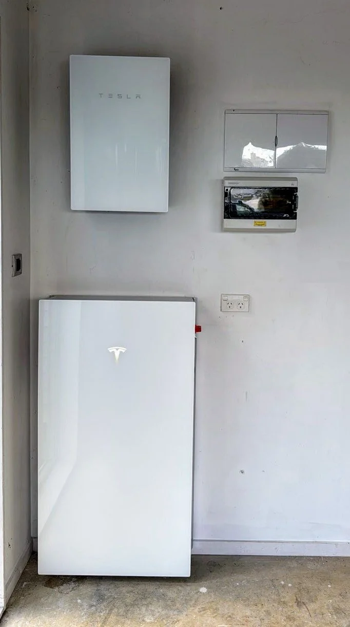 A Tesla power wall, a control panel, and a large Tesla refrigerator against a plain wall in a garage or utility room.