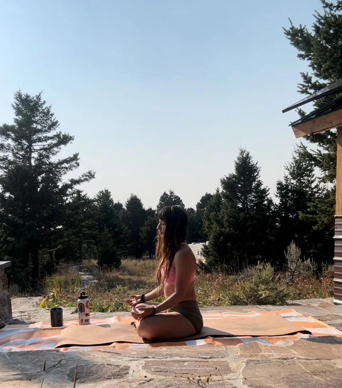 A woman practicing yoga outdoors on a mat, sitting in a cross-legged position, surrounded by trees under a clear sky.