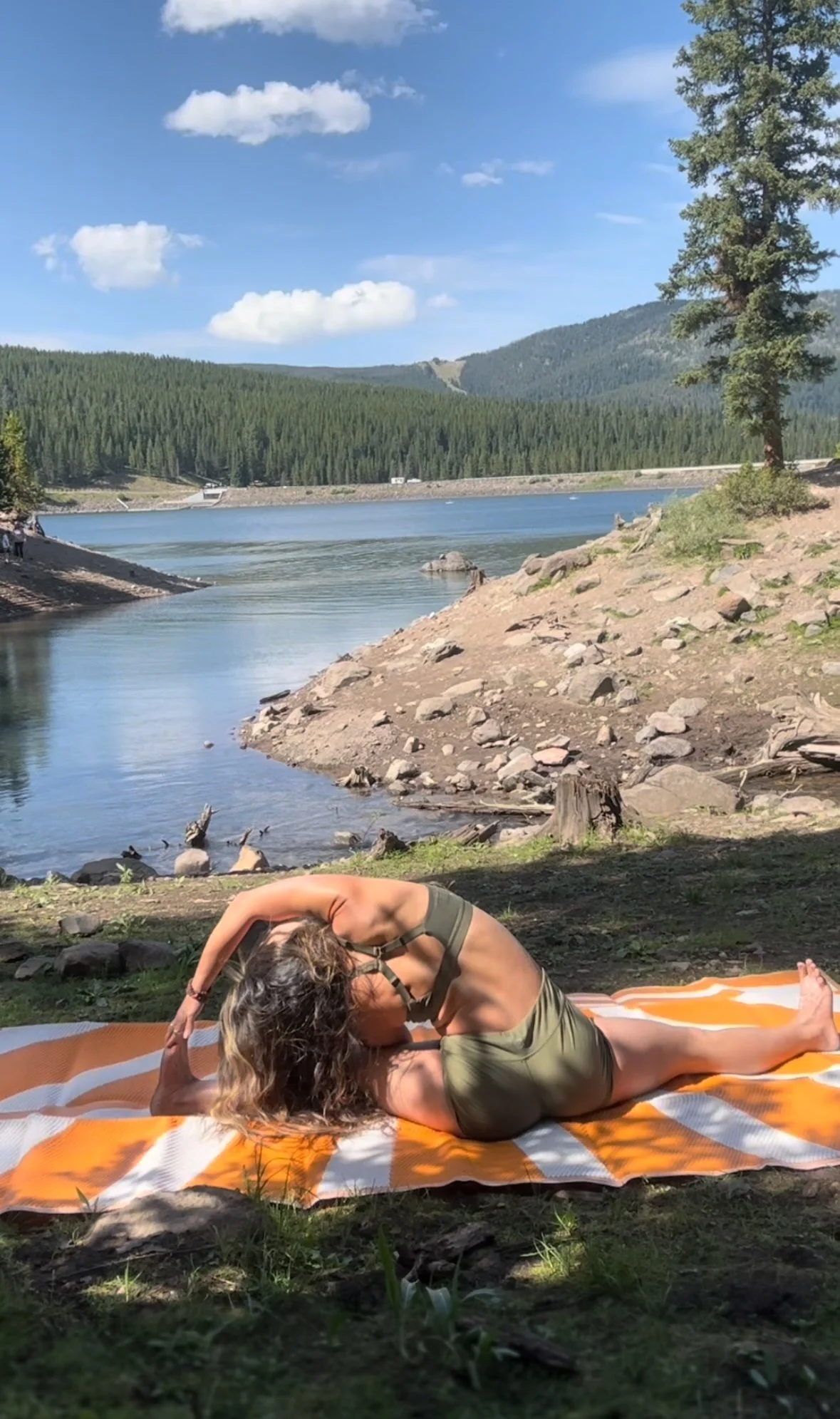 A woman practicing yoga on a striped orange and white mat by a lake, surrounded by trees, mountains, and a clear blue sky.