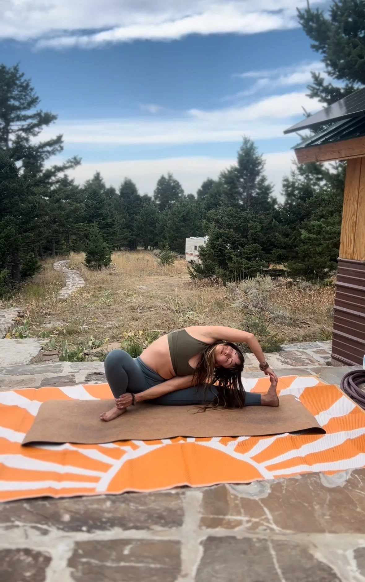 Woman practicing yoga outdoors on a stone patio, sitting on a mat with an orange and white pattern, surrounded by trees and a scenic sky.