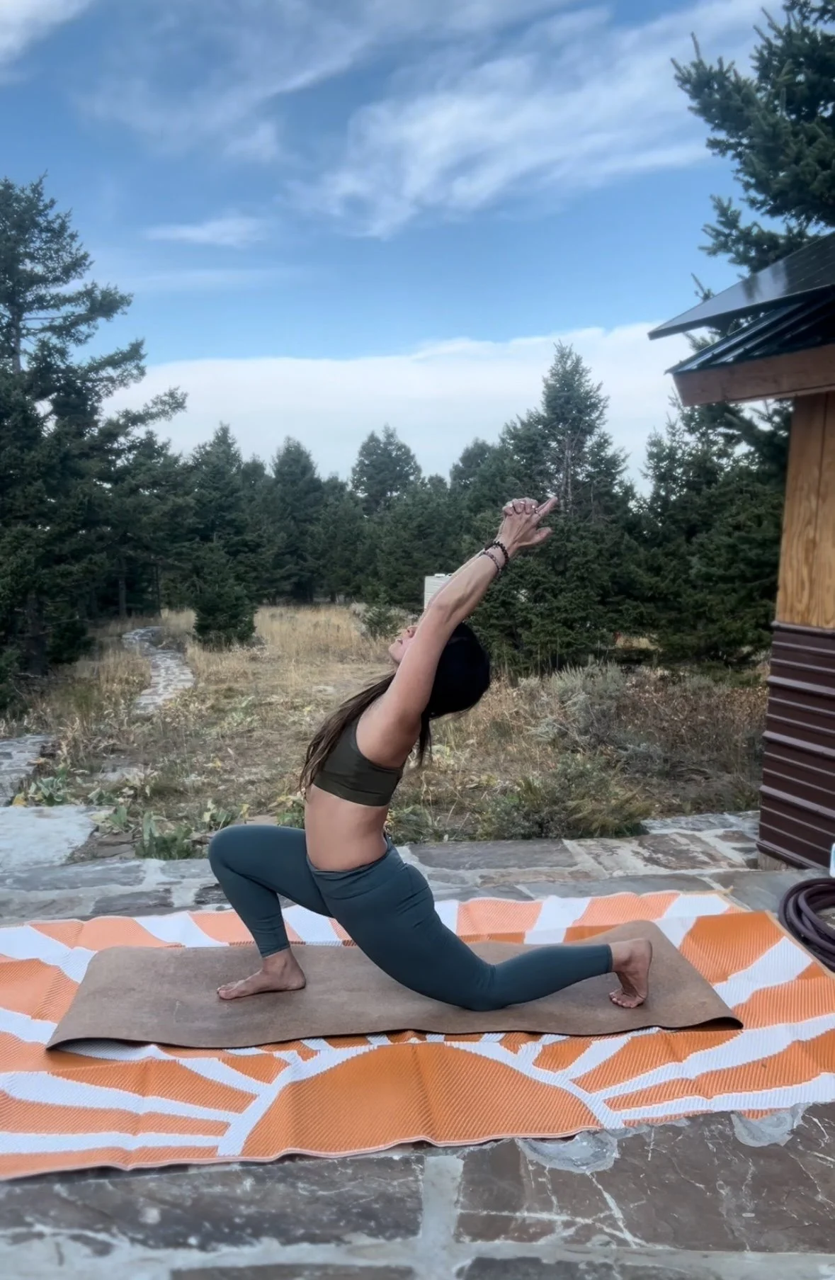 A woman practicing yoga outdoors on a mat, doing a low lunge with arms raised upwards, surrounded by trees and a cloudy sky.