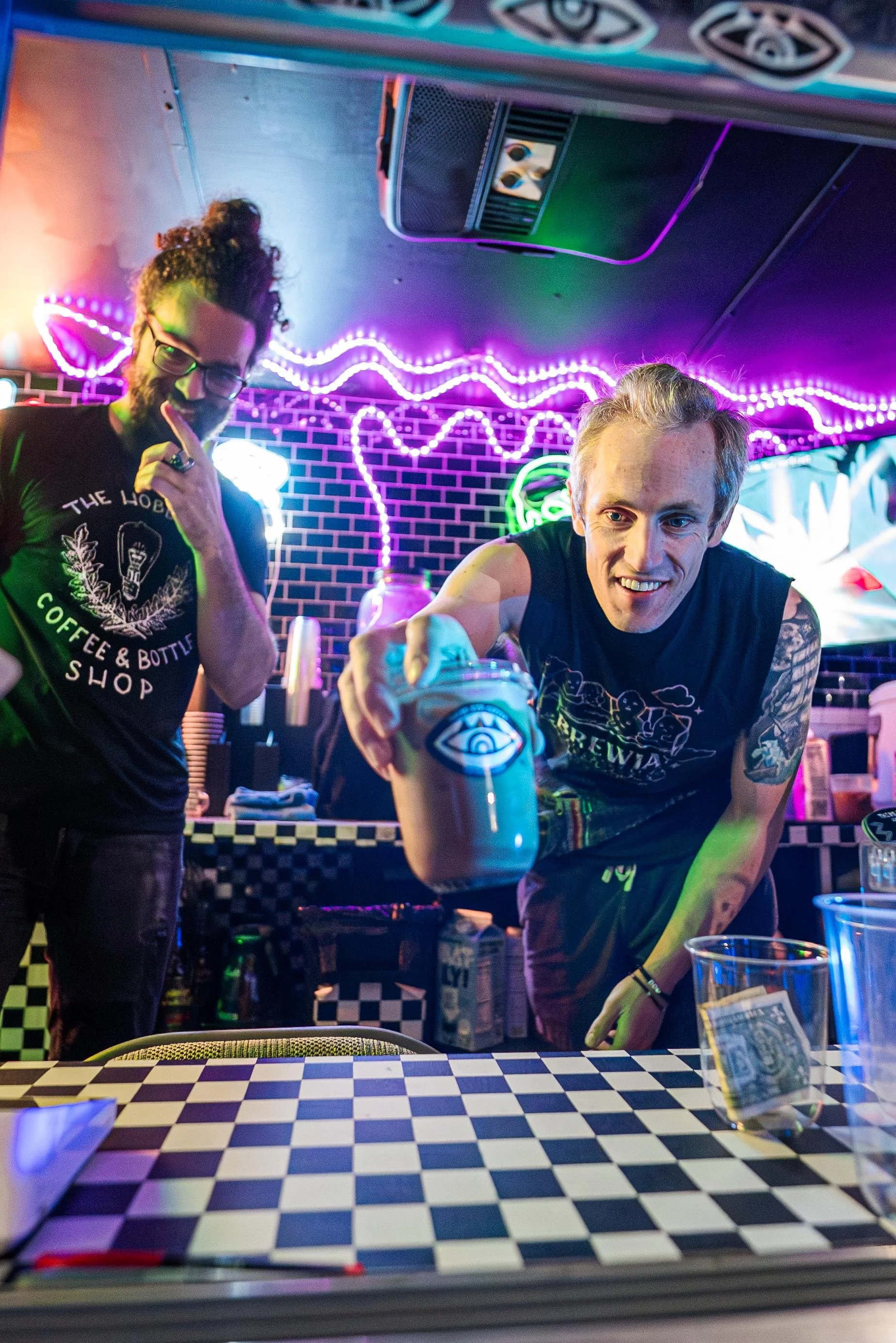 Two men behind a checkered table in a neon-lit bar, one holding a jar with a blue eye symbol, the other has a hand near his mouth, both smiling and wearing black t-shirts with designs, with neon lights and a brick wall in the background.