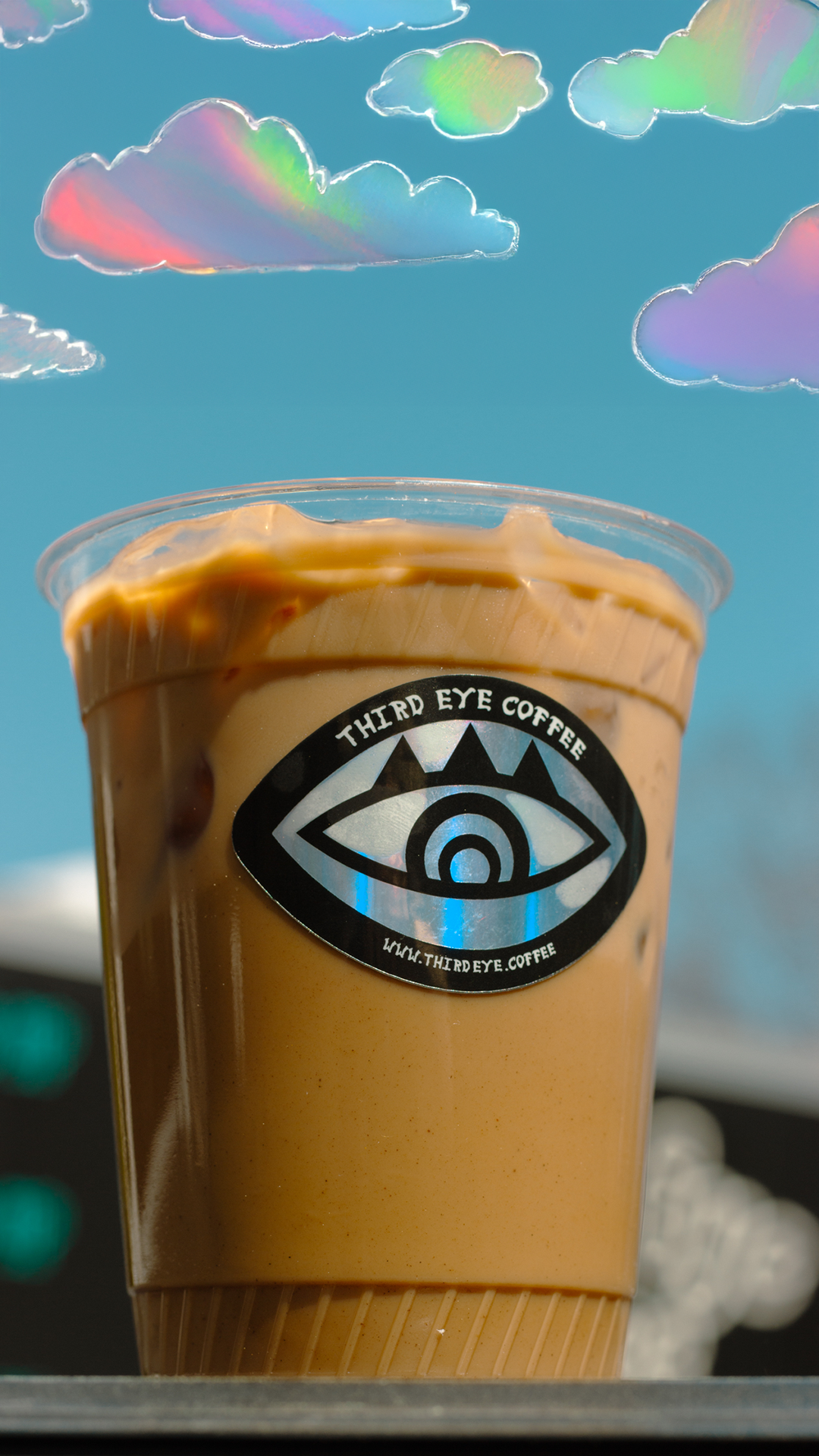 A plastic cup of iced coffee from Third Eye Coffee with a logo of an eye and the website www.thirdeye.coffee, set against a bright blue sky with digitally illustrated pastel-colored clouds.