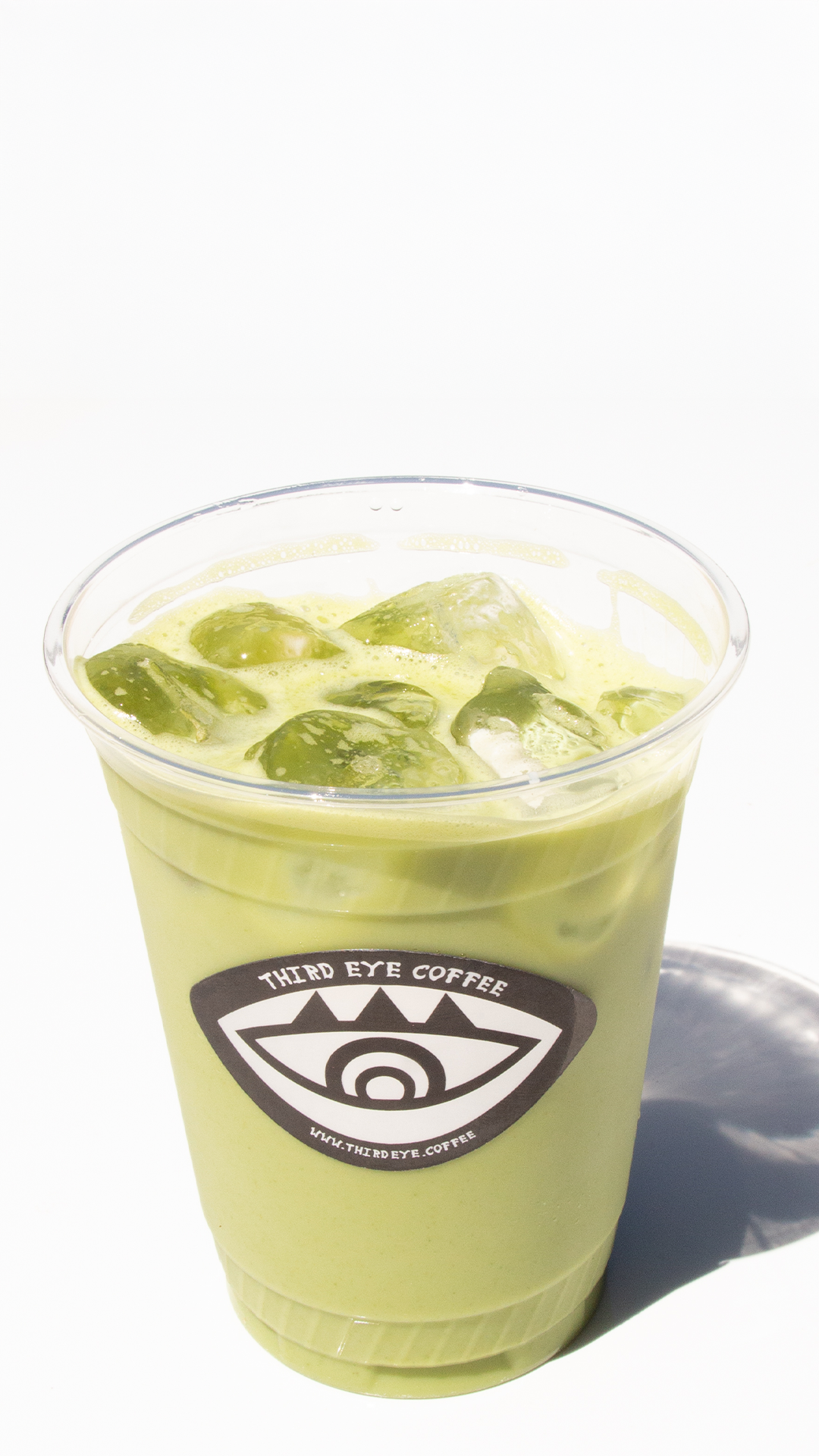 A clear plastic cup filled with green matcha latte and ice cubes, bearing a 'Third Eye Coffee' logo with a stylized eye design.
