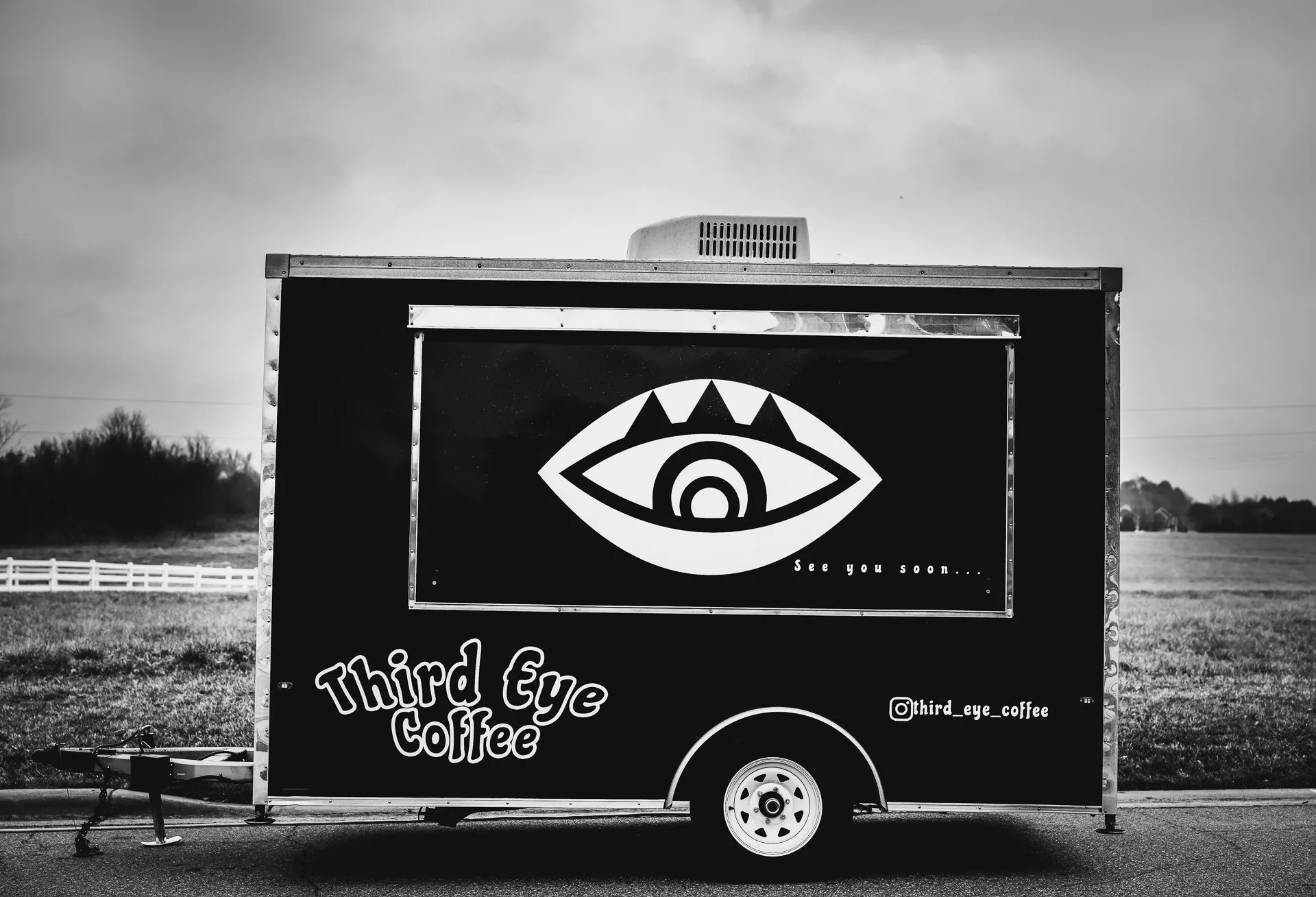 A black food truck with a large eye graphic on the side and the text "Third Eye Coffee" and social media handle @third_eye_coffee. The background shows an open field with trees and a cloudy sky.