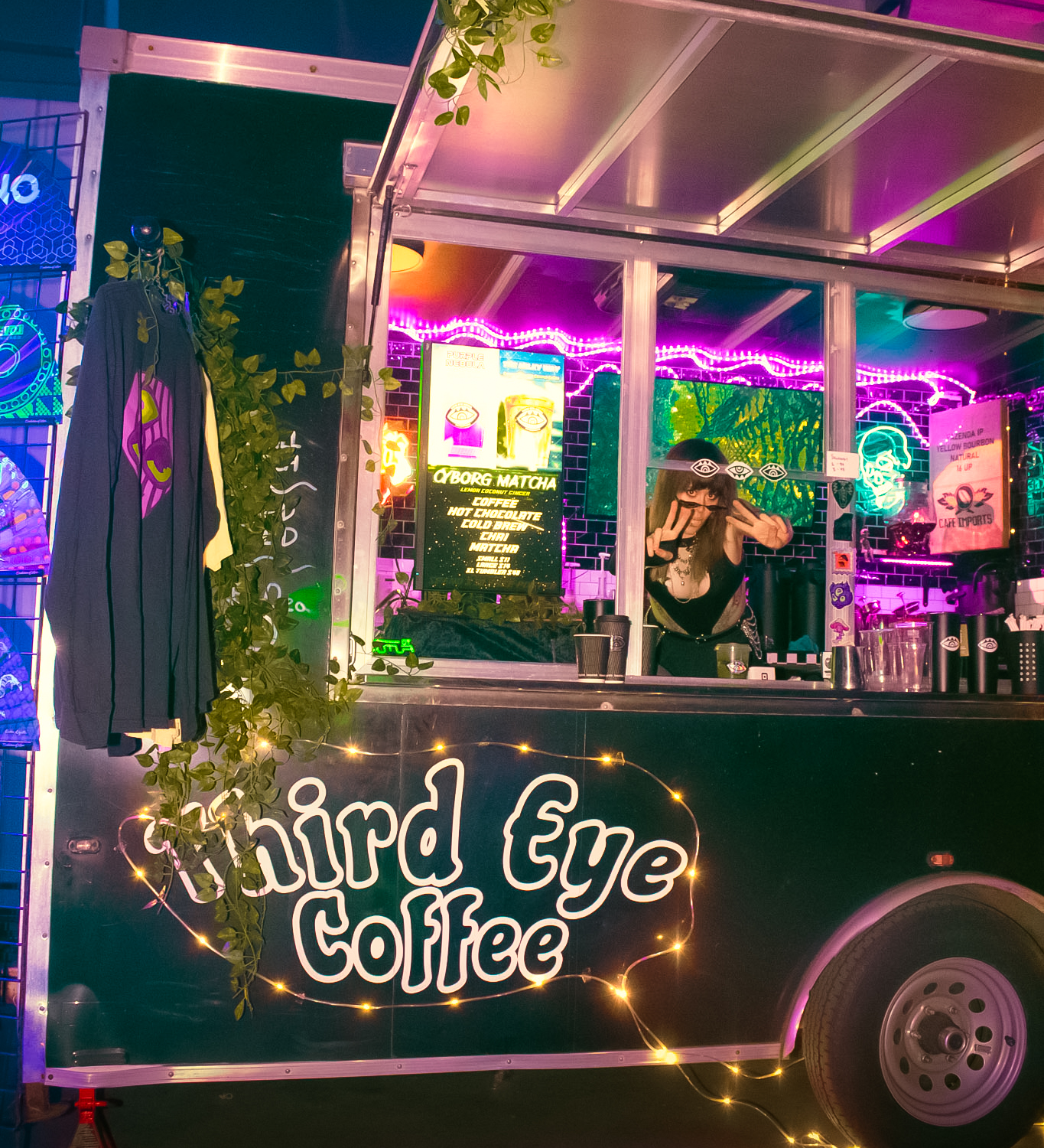 A black food truck named 'Chirid Eye Coffee' decorated with string lights and greenery, serving coffee and drinks at night with neon signs and a woman making a peace sign inside.